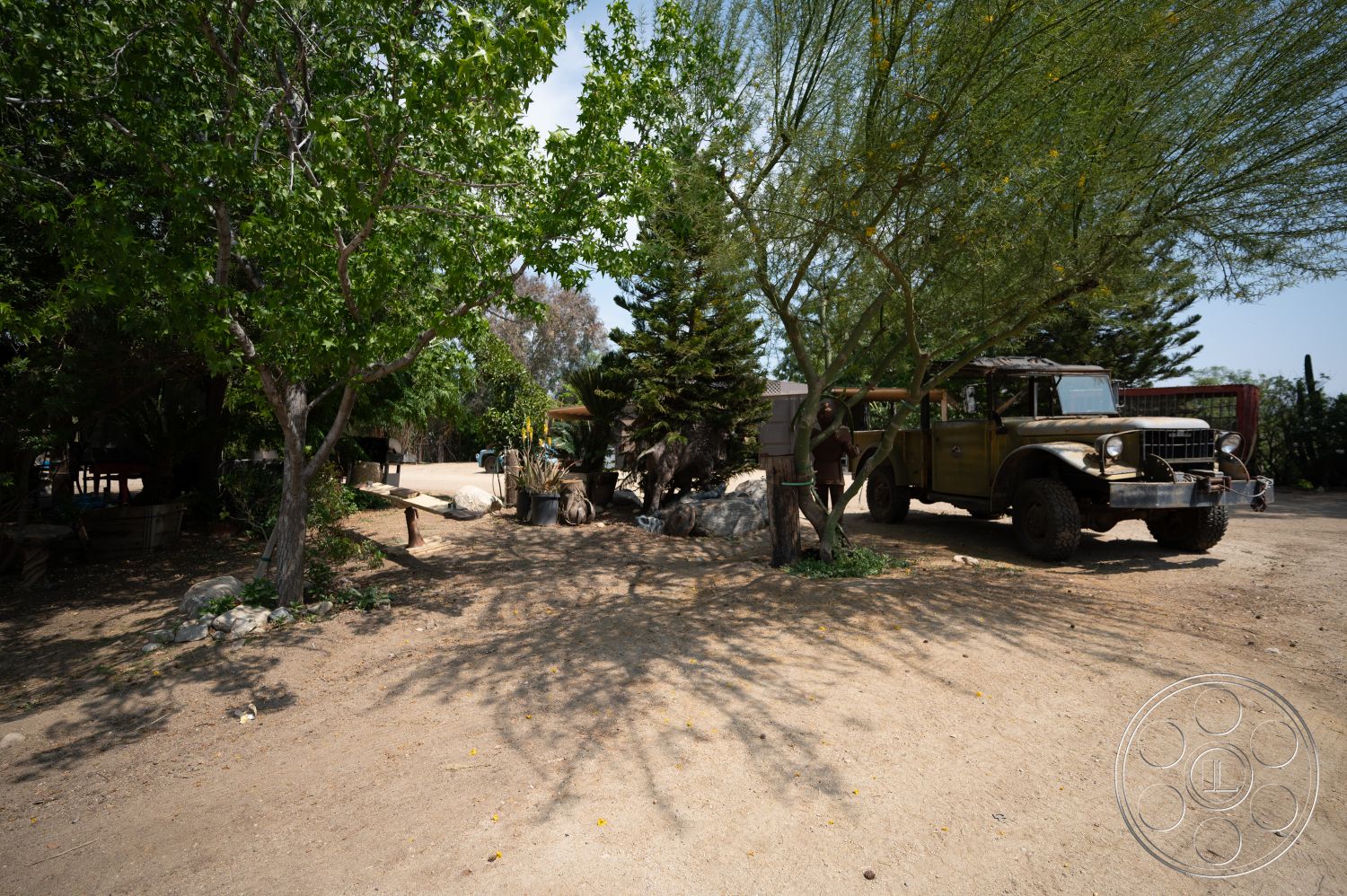 Ranch 6 - exterior, natural stone elements, agave plants, dirt driveway, desert setting, rock landscaping, lush green trees, rustic outdoor furniture, large outdoor vehicle, shadowed areas from trees