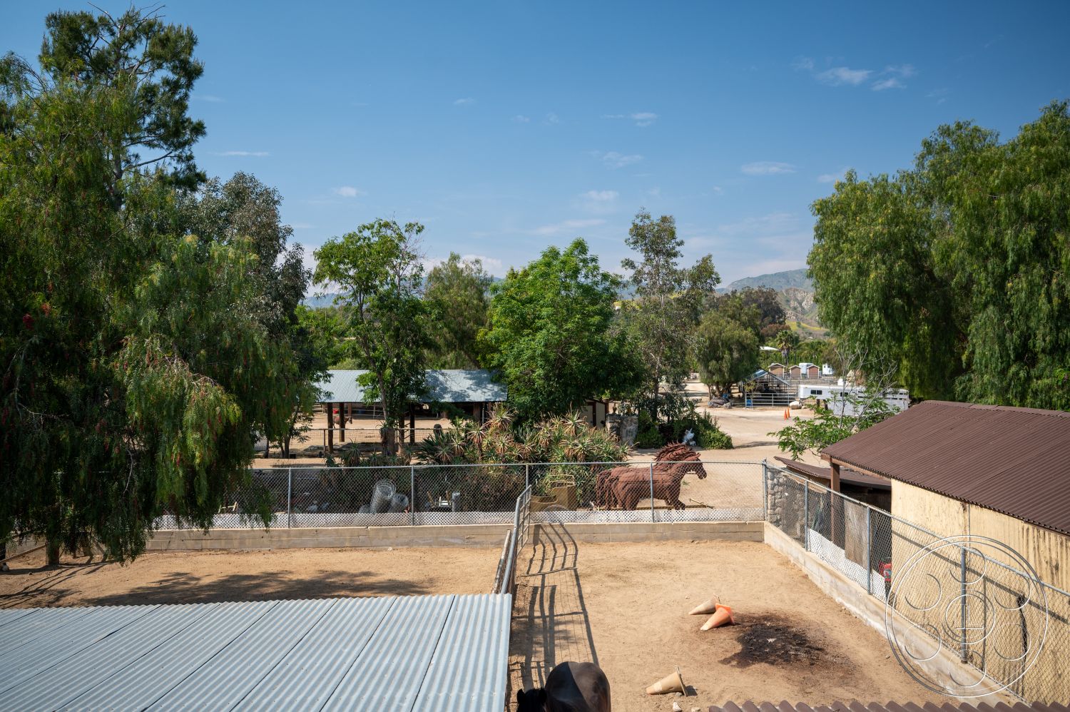Ranch 6 - exterior, metal roofing, flat terrain, wooden barn structure, agricultural environment, chain-link fence enclosure, dirt paddock area, landscape with trees, sculpted horse art, outdoor ranch setting, open space pasture