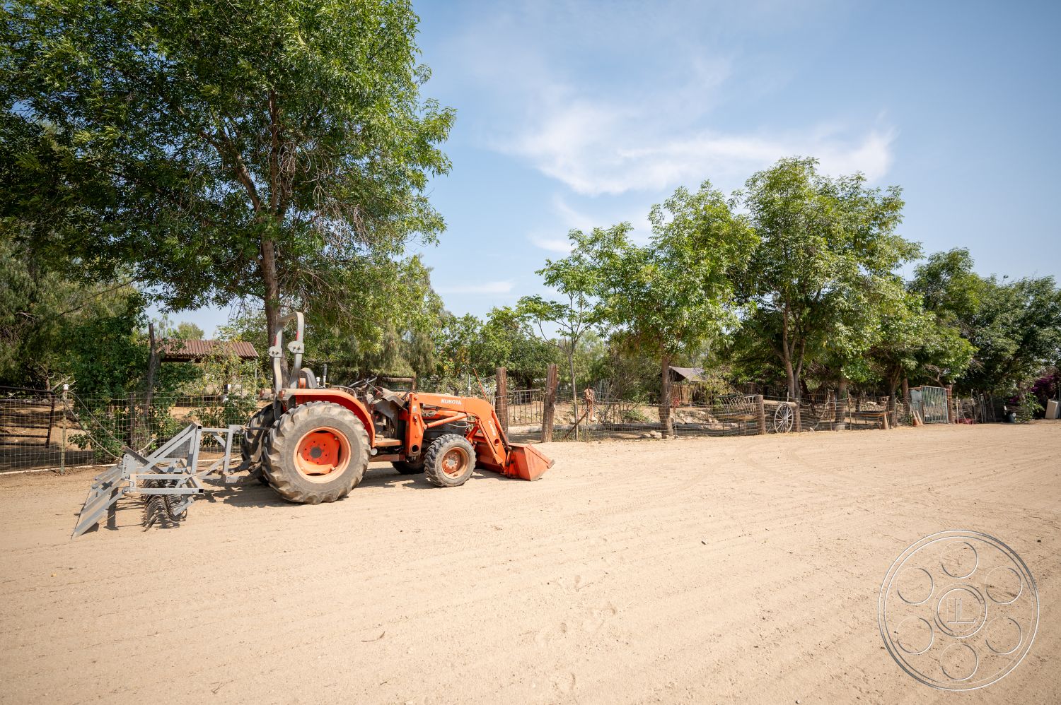 Ranch 6 - exterior, clear blue sky, wooden fence, agricultural land, green leafy trees, sunny day, open field setting, agricultural equipment, dirt farm path, rustic tractor with front loader, farmyard environment