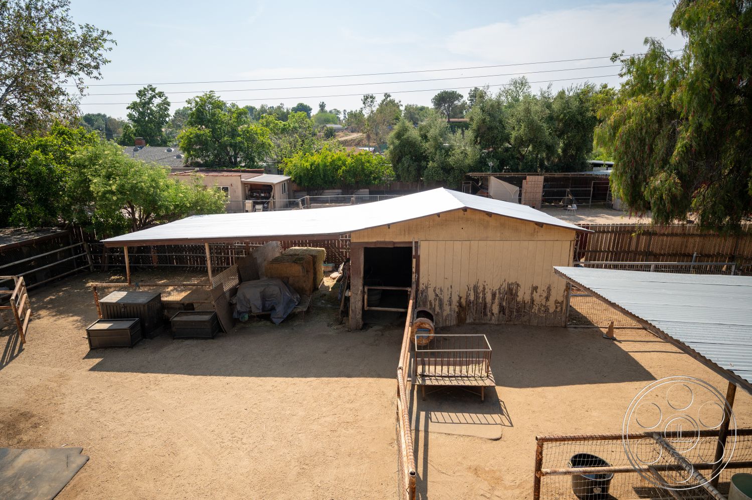Ranch 6 - exterior, open outdoor space, metal roof, surrounding trees and greenery, wooden barn structure, dirt ground surface, fenced animal enclosure, hay storage area, wooden post and rail fence
