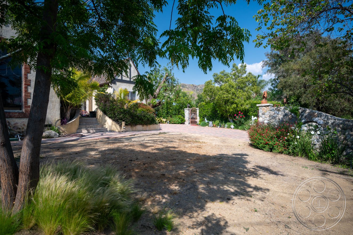 Ranch 6 - exterior, lush greenery, palm trees, paver driveway, brick steps, flowering plants, desert landscape, arched entryway, wooden railing, two-story home, landscaped garden area, stucco exterior walls, stone garden wall