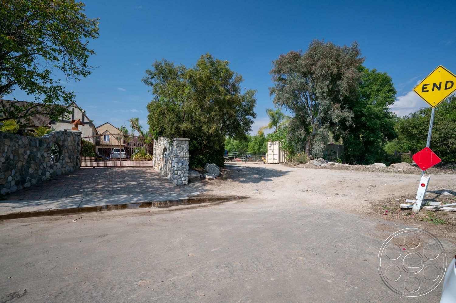 Ranch 6 - iron gate, gravel driveway, natural landscape setting, brick driveway pavers, sunny day, open outdoor area, exterior stone wall entrance, tall green trees, residential property access, end of road sign