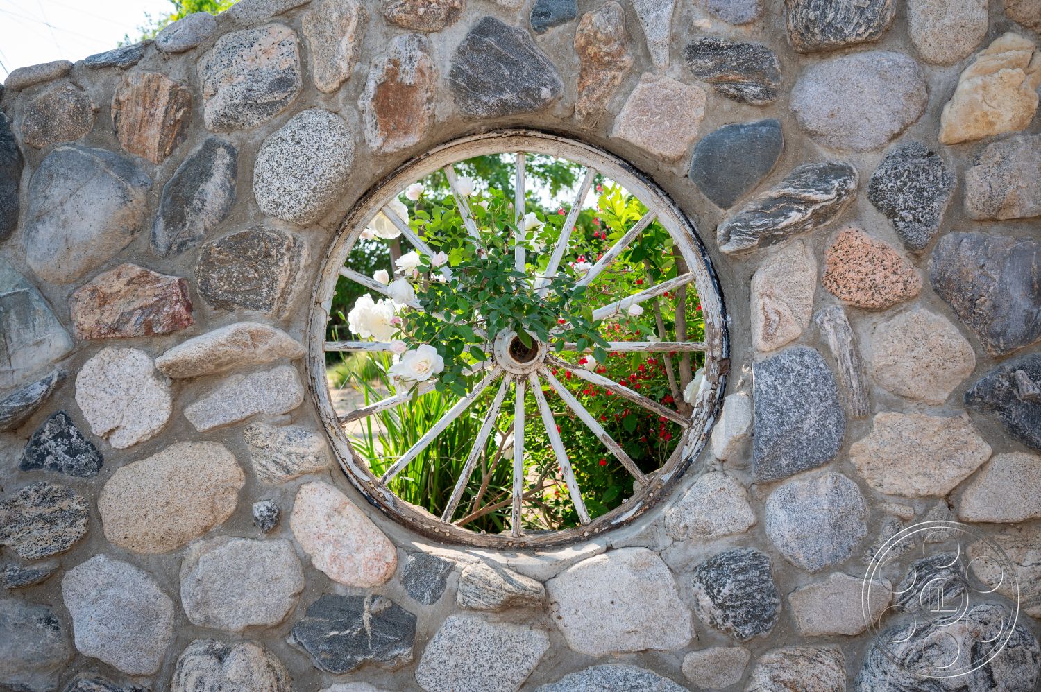 Ranch 6 - landscaped garden view, natural stone exterior, stone wall construction, circular stone window opening, circular wood wheel feature, bright floral accents, exterior wall treatment