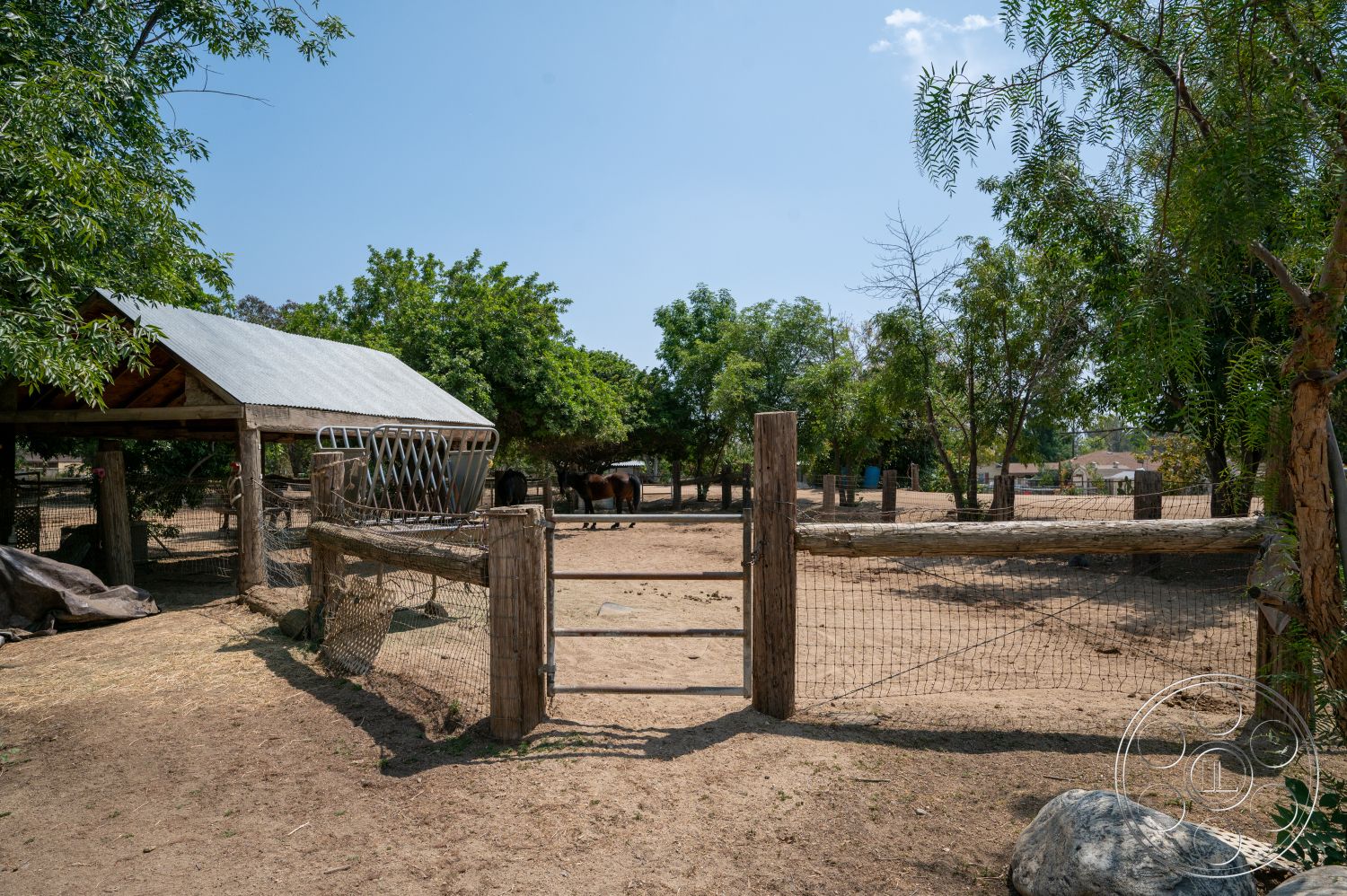 Ranch 6 - exterior, open outdoor space, farm setting, gravel ground surface, sunny rural environment, wooden corral fencing, weathered wooden posts, metal wire mesh, thatched roof animal shelter, deciduous tree landscape, dirt path access