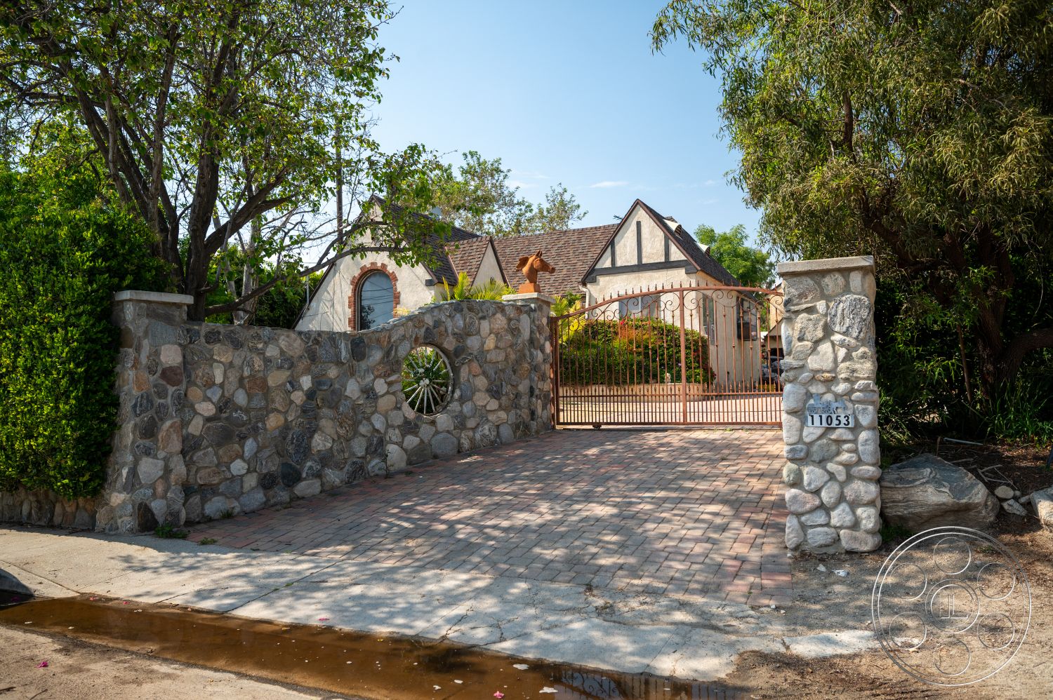 Ranch 6 - exterior, landscaped garden, mature trees, stucco walls, arched entryway, stone driveway, arched windows, gable roof, wrought iron gate, brick paver driveway, stone gate pillars, siding walls