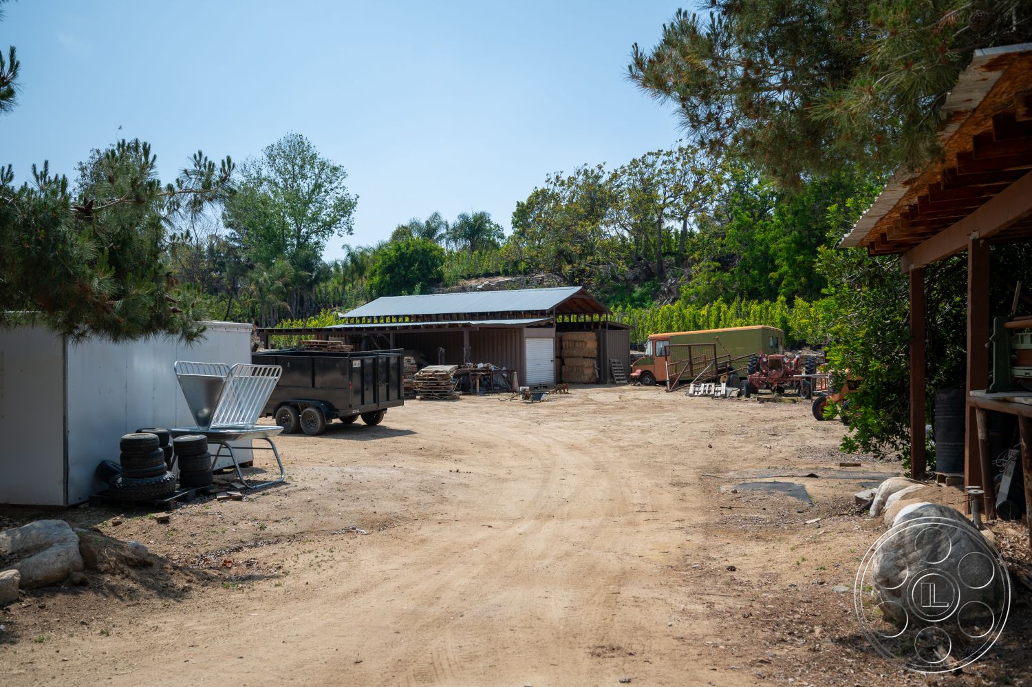 Ranch 6 - exterior, rural setting, gravel driveway, dirt ground, open outdoor area, wooden barn structure, metal storage container, rusted farm equipment, weathered wooden fence, tin roof, tree line background