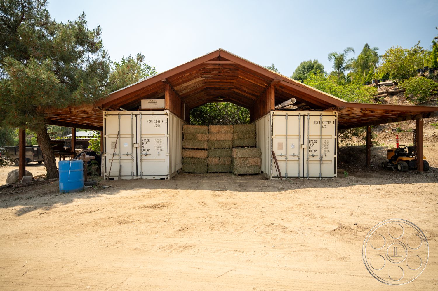 Ranch 6 - exterior, outdoor equipment, rural setting, tree landscaping, supporting columns, recessed lighting fixtures, natural vegetation backdrop, dirt ground surface, open-air storage space, wooden roof structure, metal shipping containers, stacked hay bales, blue plastic drum