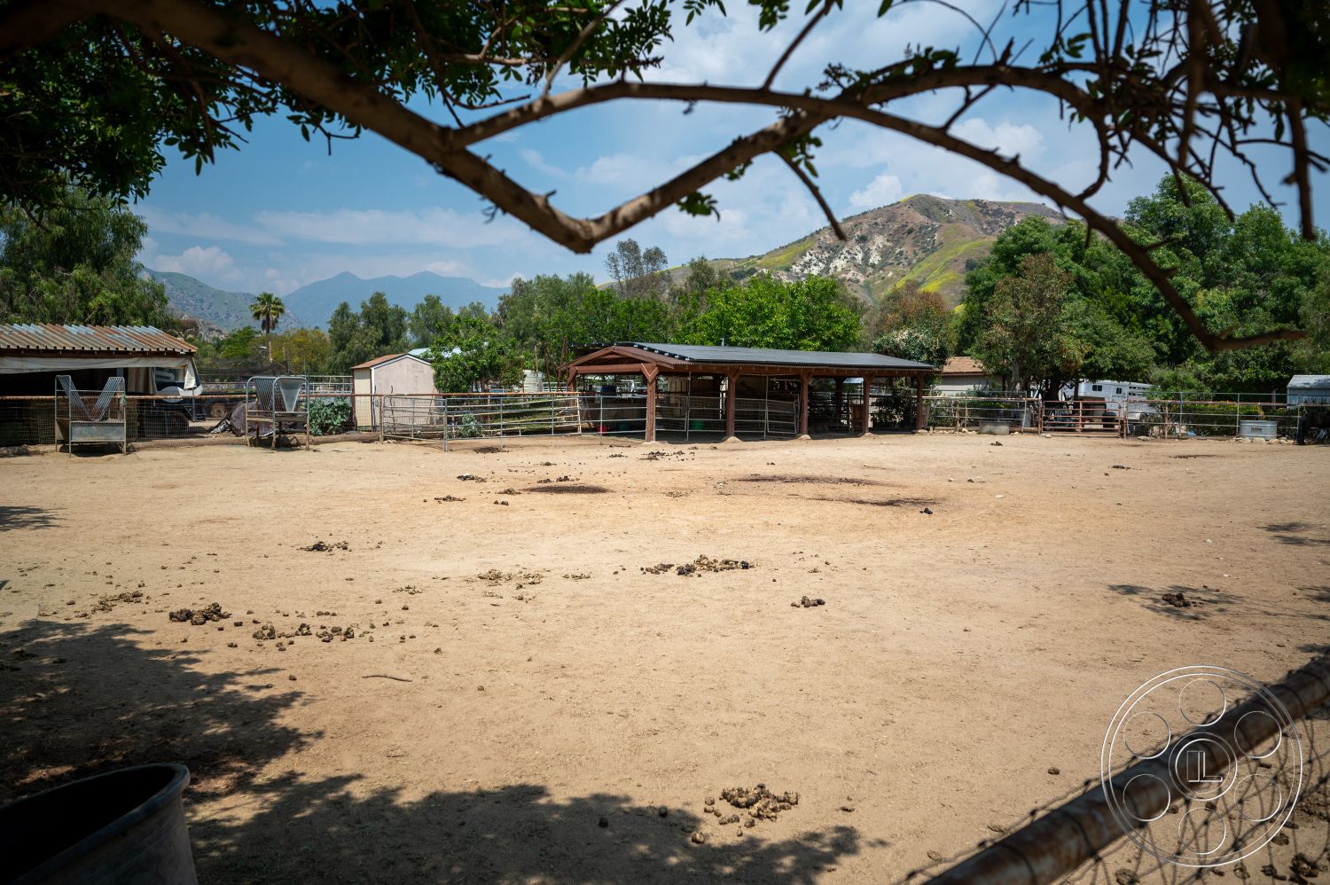 Ranch 6 - exterior, surrounding trees, clear blue sky, metal roof, wooden barn structure, dirt paddock area, fenced livestock enclosure, mountainous background landscape