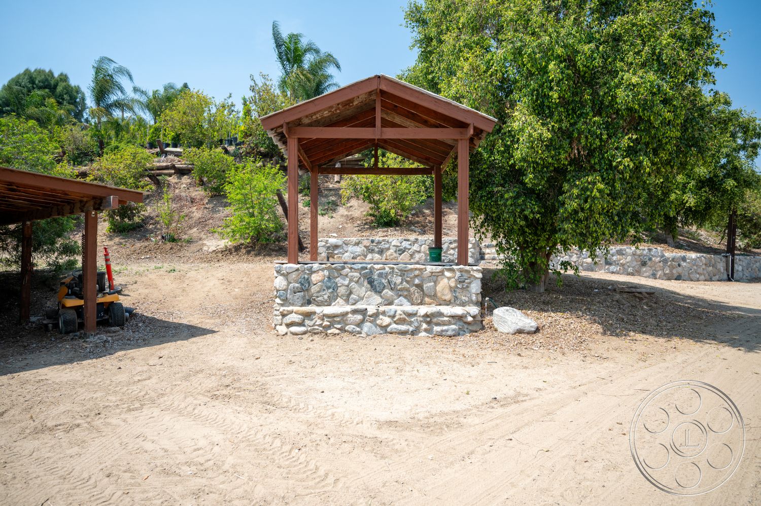 Ranch 6 - exterior, exposed wooden beams, tropical palm trees, dirt pathway, gravel landscape, drought-resistant plants, vegetation border, natural hillside setting, stone gazebo base, wooden gazebo roof