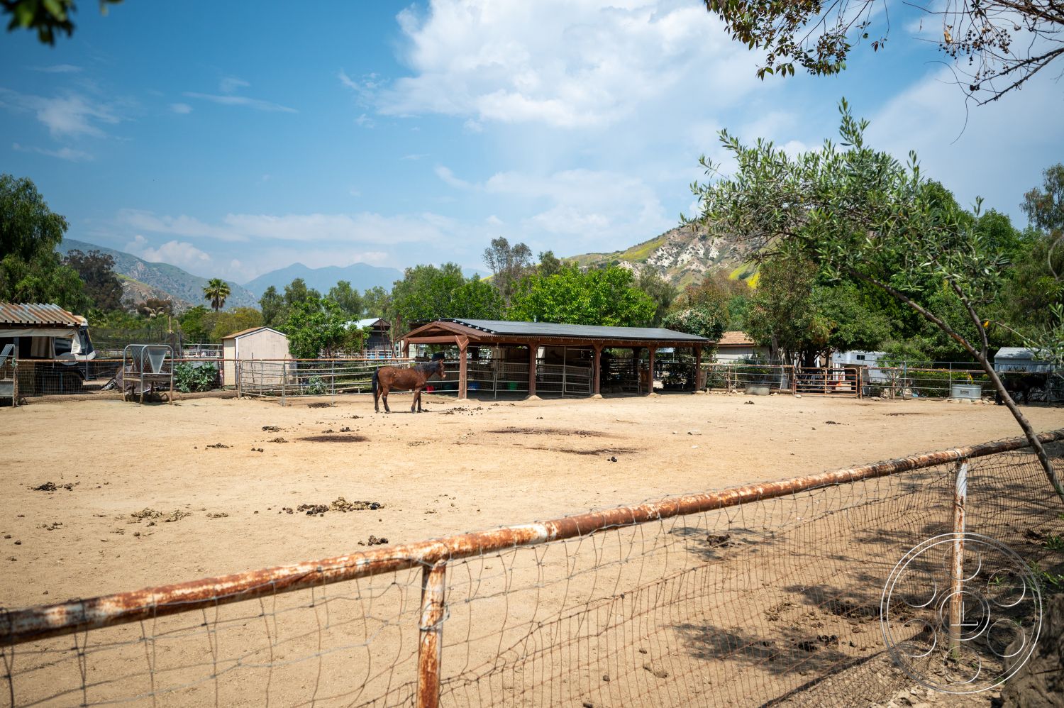 Ranch 6 - exterior, wooden fence, lush greenery surrounding, mountain landscape backdrop, dirt paddock area, wooden horse barn, metal roof on barn, animal shelter structures, outdoor space for horses, country ranch setting