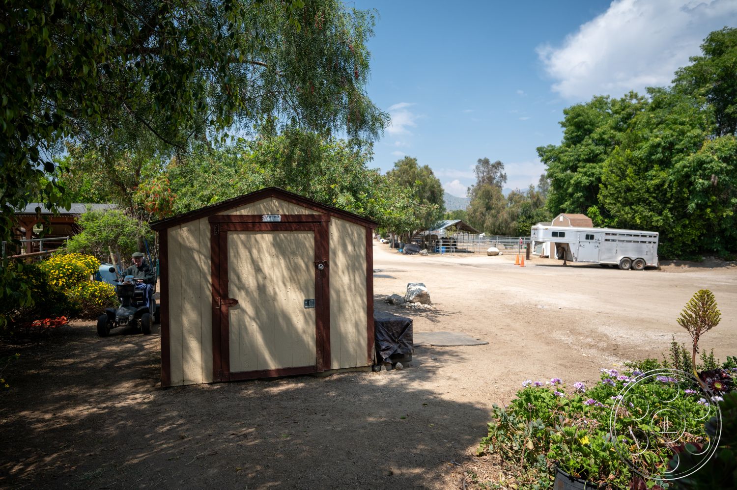 Ranch 6 - exterior, lush greenery, gravel pathway, mountain backdrop, metal roof, dirt parking area, outdoor environment, agricultural setting, landscape with flowering plants, brown trim details, farm equipment, wood storage shed, cream-colored siding, freestanding trailer