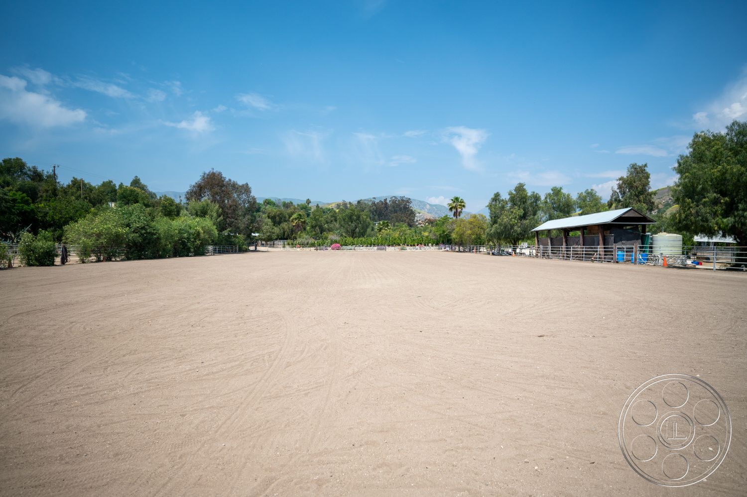 Ranch 6 - exterior, open outdoor space, mountain backdrop, blue sky, landscaped with trees, wooden fencing, sunny weather, dirt riding arena surface, green vegetation surrounding area, metal roof on barn structure