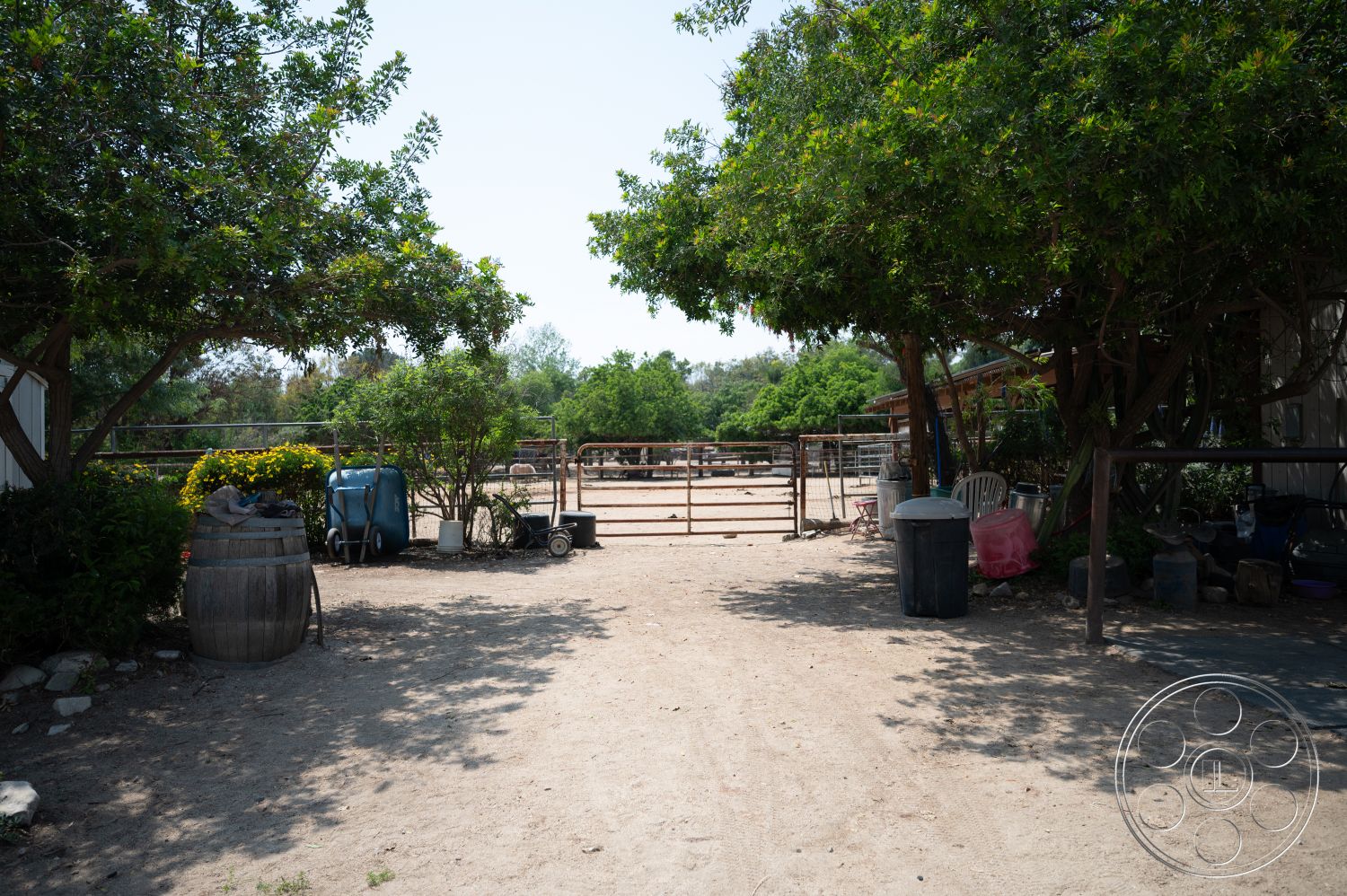 Ranch 6 - exterior, outdoor seating area, dirt pathway, wooden fencing, lush green foliage, landscaped garden beds, sunny rural setting, horse corral area, wooden barrel planter, metal trash bins