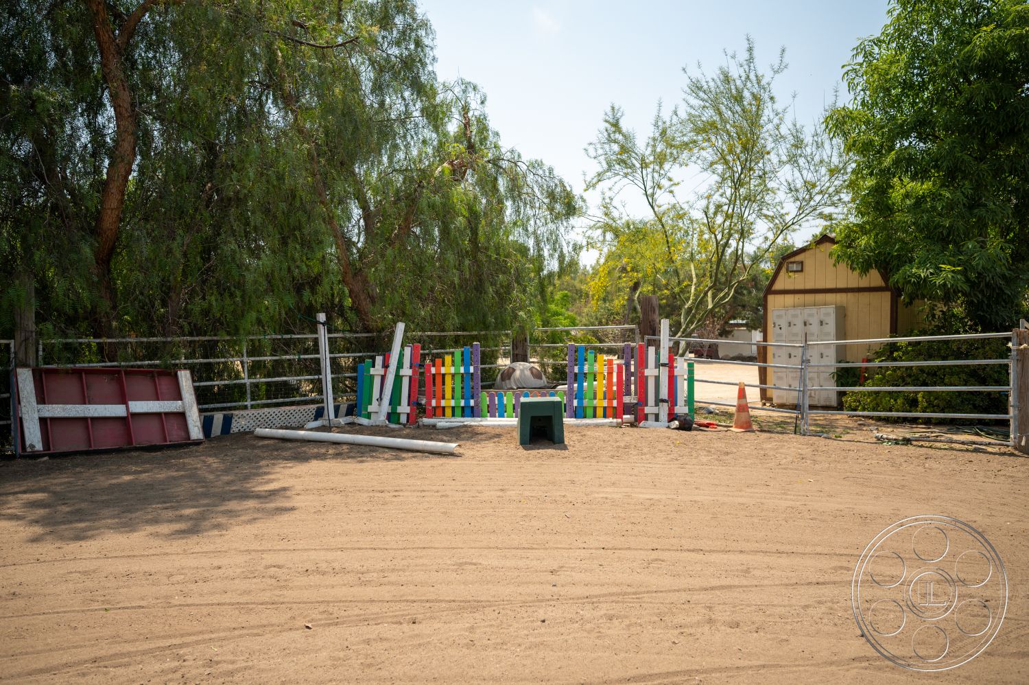 Ranch 6 - exterior, lush vegetation, gravel driveway, wooden fencing, sunny rural setting, rustic storage shed, sand arena floor, colorful wooden jump poles, metal horse corral panels, outdoor riding area