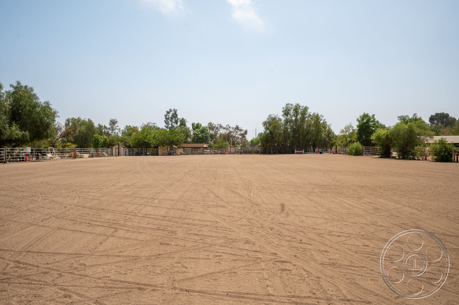 Ranch 6 - open space, clear blue sky, rural setting, sand surface, fenced perimeter, sunny outdoor environment, tree-lined border, dirt riding arena