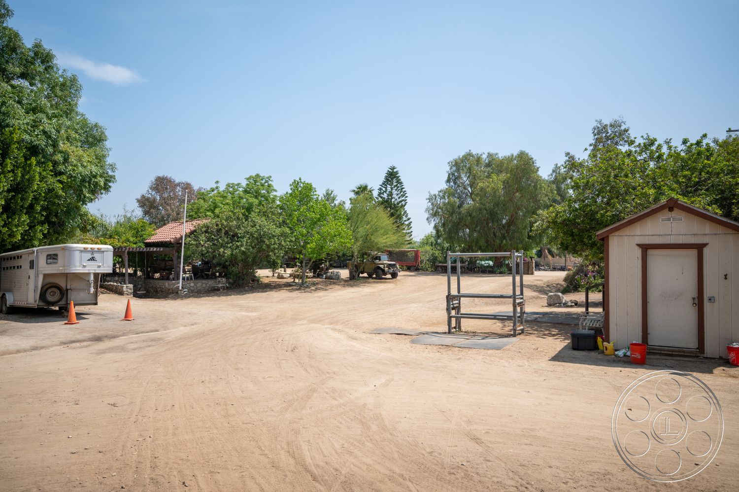 Ranch 6 - exterior, metal fencing, rural setting, dirt driveway, open landscape, horse trailer, wooden shed with metal roof, covered outdoor area with stone pillars, green foliage with trees and bushes, parking cones