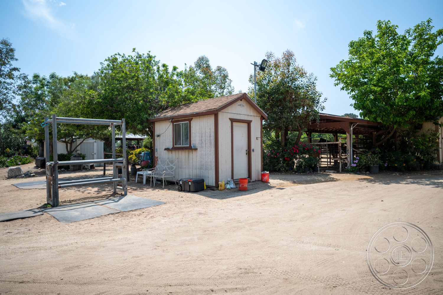 Ranch 6 - green foliage, dirt driveway, metal light fixture, landscaped garden area, shaded outdoor seating, rustic farm setting, wood shed exterior, wood framed door, concrete base pads, metal poles, graveled surroundings, wooden support structure