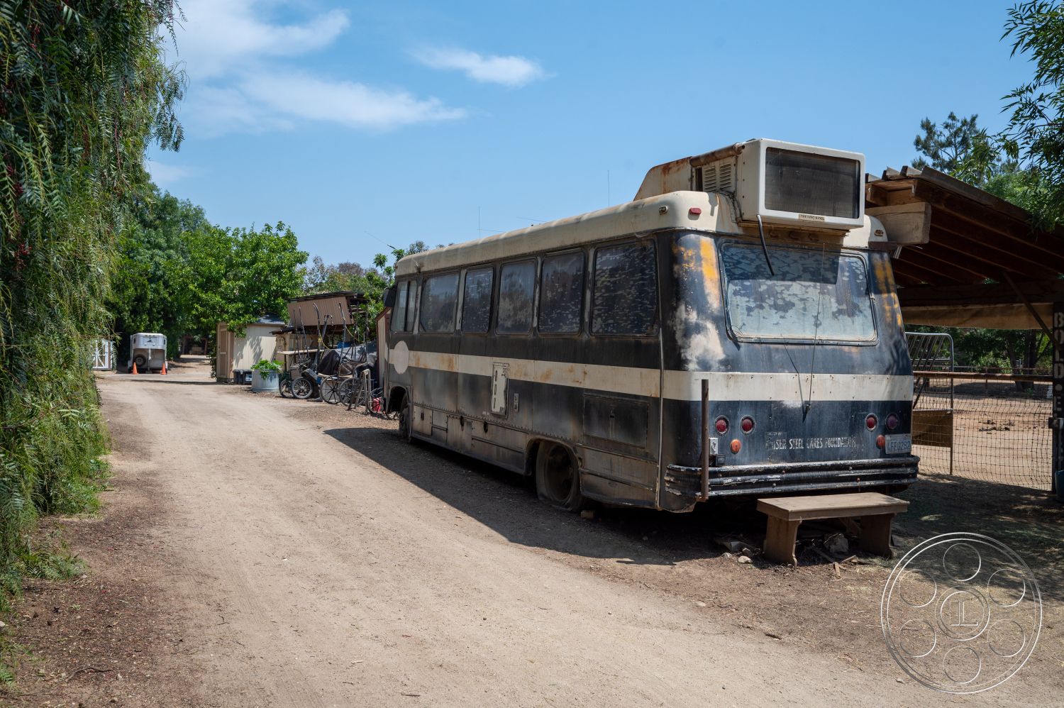 Ranch 6 - exterior, surrounding greenery, open outdoor space, dirt pathway, overhanging tree canopy, rusted bus exterior, large windows on bus, outdoor air conditioning unit, bicycle leaning against bus, wooden bench next to bus, rustic shed structure, fenced area nearby, casual rural setting
