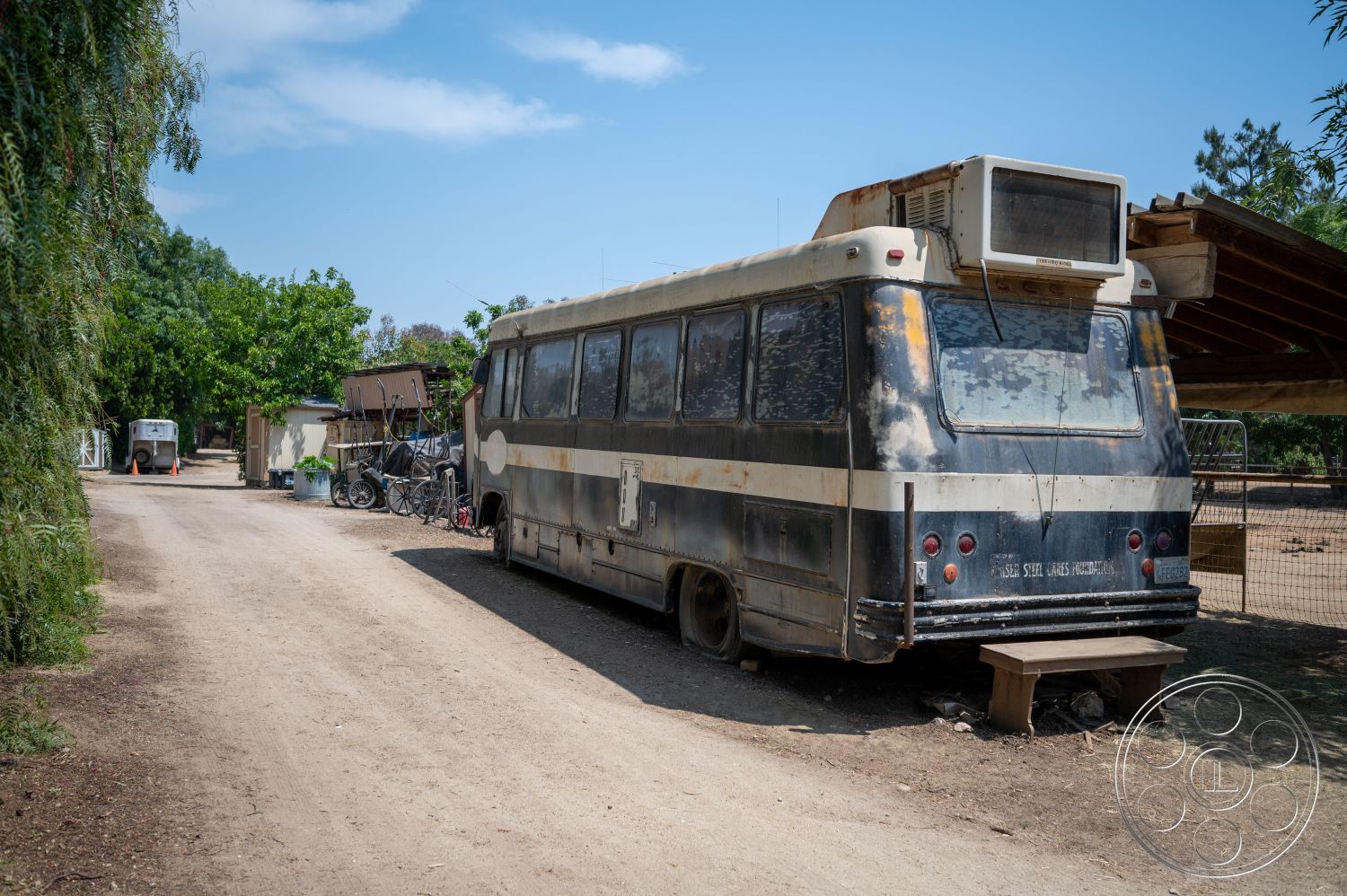 Ranch 6 - exterior, surrounding greenery, gravel path, dirt driveway, open sky view, rusted metal bus exterior, weathered wood overhang, colorful bicycles, vintage style bus