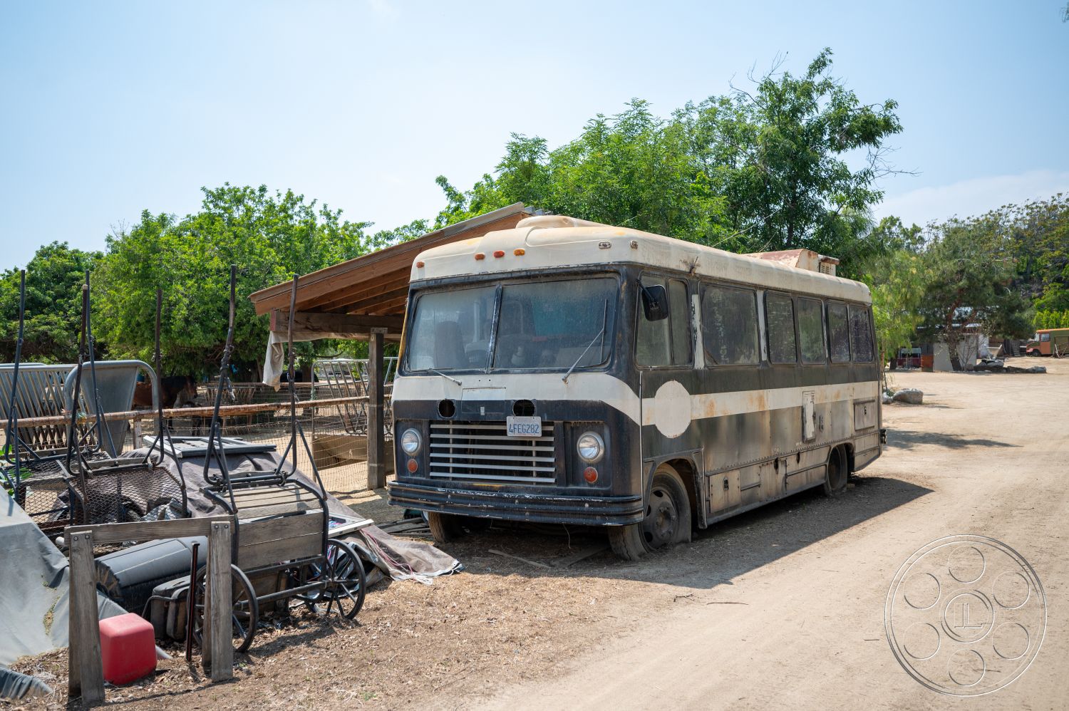 Ranch 6 - exterior, overgrown vegetation, dirt driveway, farm setting, sunny outdoor environment, weathered metal siding, vintage bus, wooden structure awning, rusted metal cart, abandoned landscape