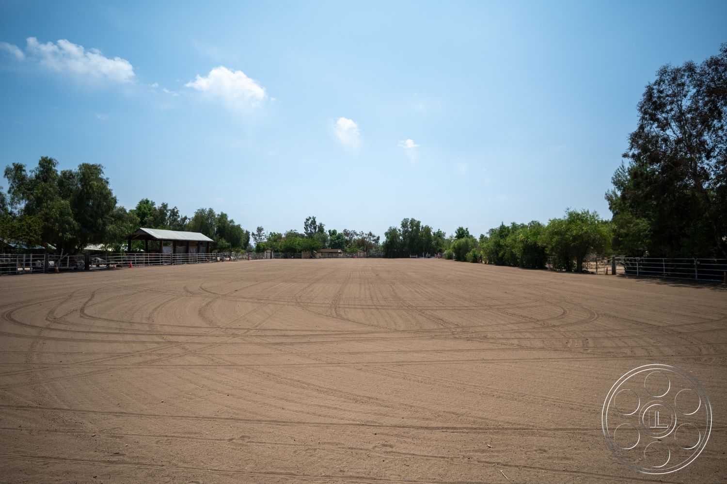 Ranch 6 - metal fencing, clear blue sky, sunny setting, sand surface, rural environment, tree line backdrop, wooden barn structure, outdoor arena