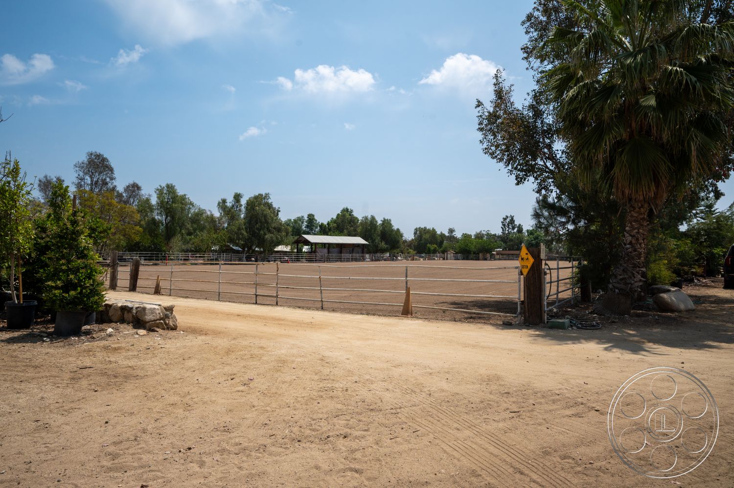 Ranch 6 - exterior, paved pathway, wooden fence, dirt driveway, grass landscaping, open sky view, suburban ranch setting, fenced equestrian area, outdoor riding arena, single-story barn structure, mixed tree landscaping, horse riding environment