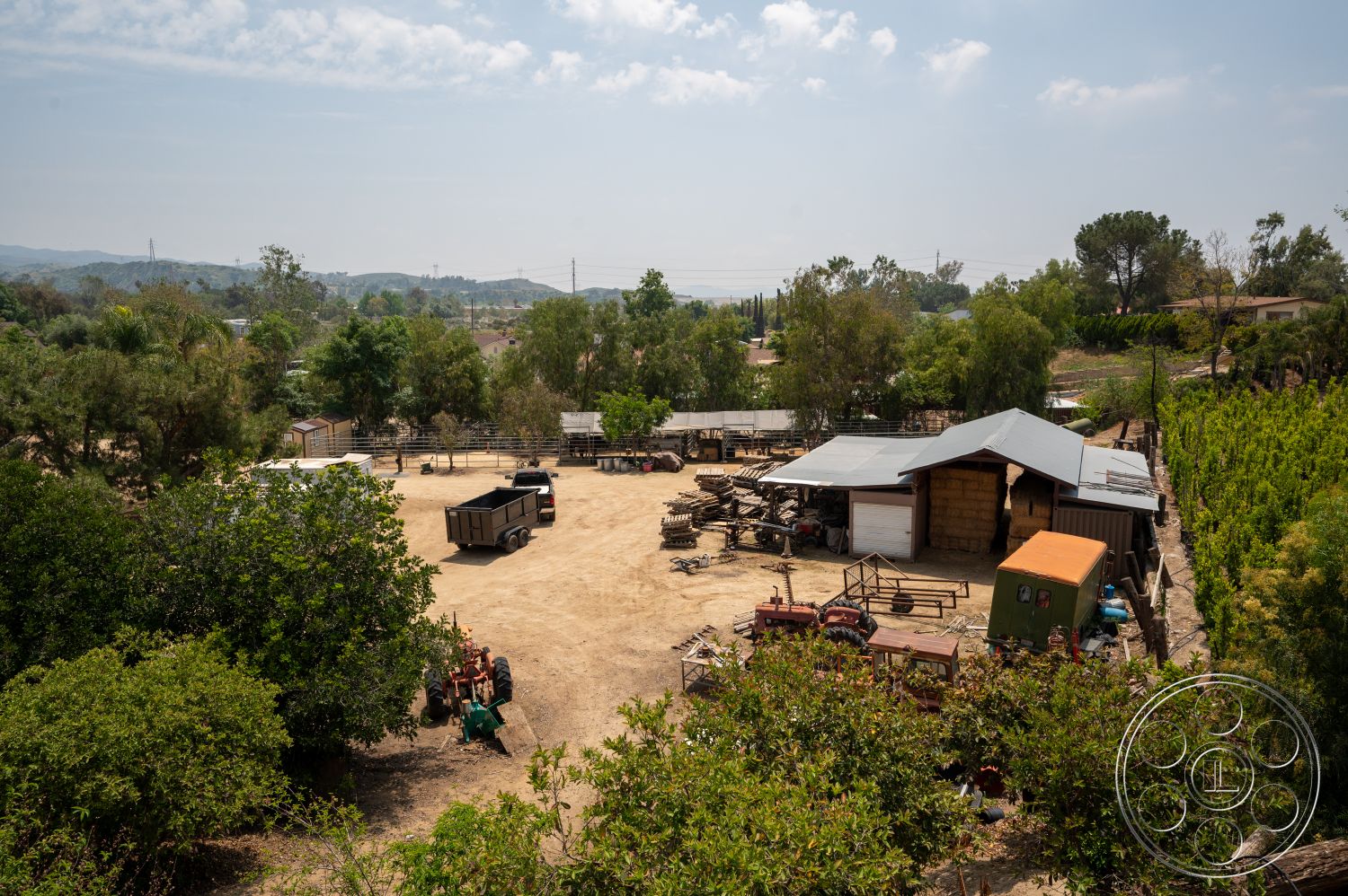 Ranch 6 - exterior, green shrubbery, asphalt driveway, wooden fence, agricultural landscape, storage shed, wooden barn structure, open pasture area, metal barn roof, farmyard setting, dirt lot surface, scattered farm equipment, rolling hills in background