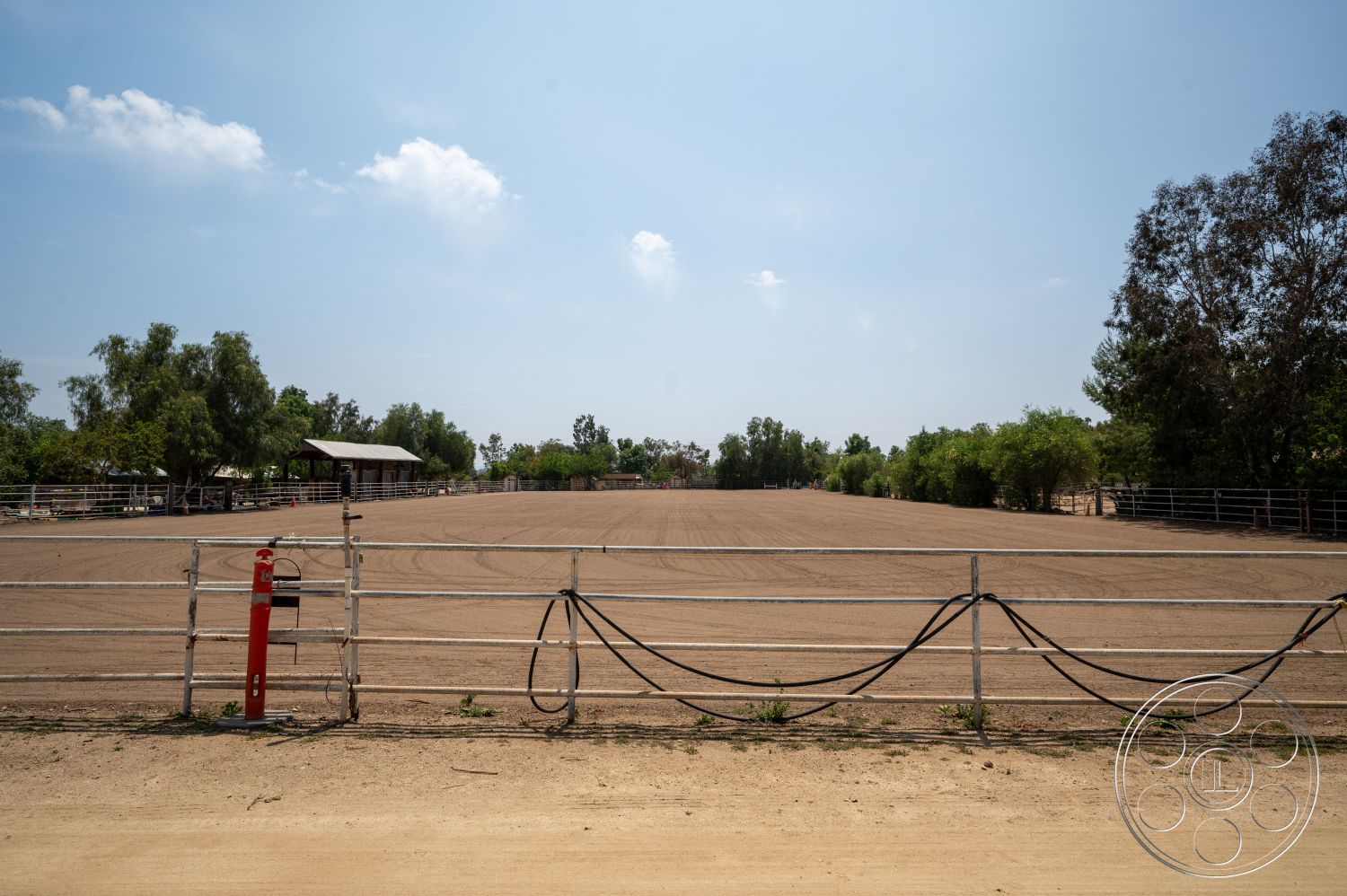 Ranch 6 - surrounding trees, clear blue sky, rural setting, wooden fence enclosure, outdoor recreational area, dirt riding arena, open pasture space, metal rail fencing, landscape with mounted shade structure