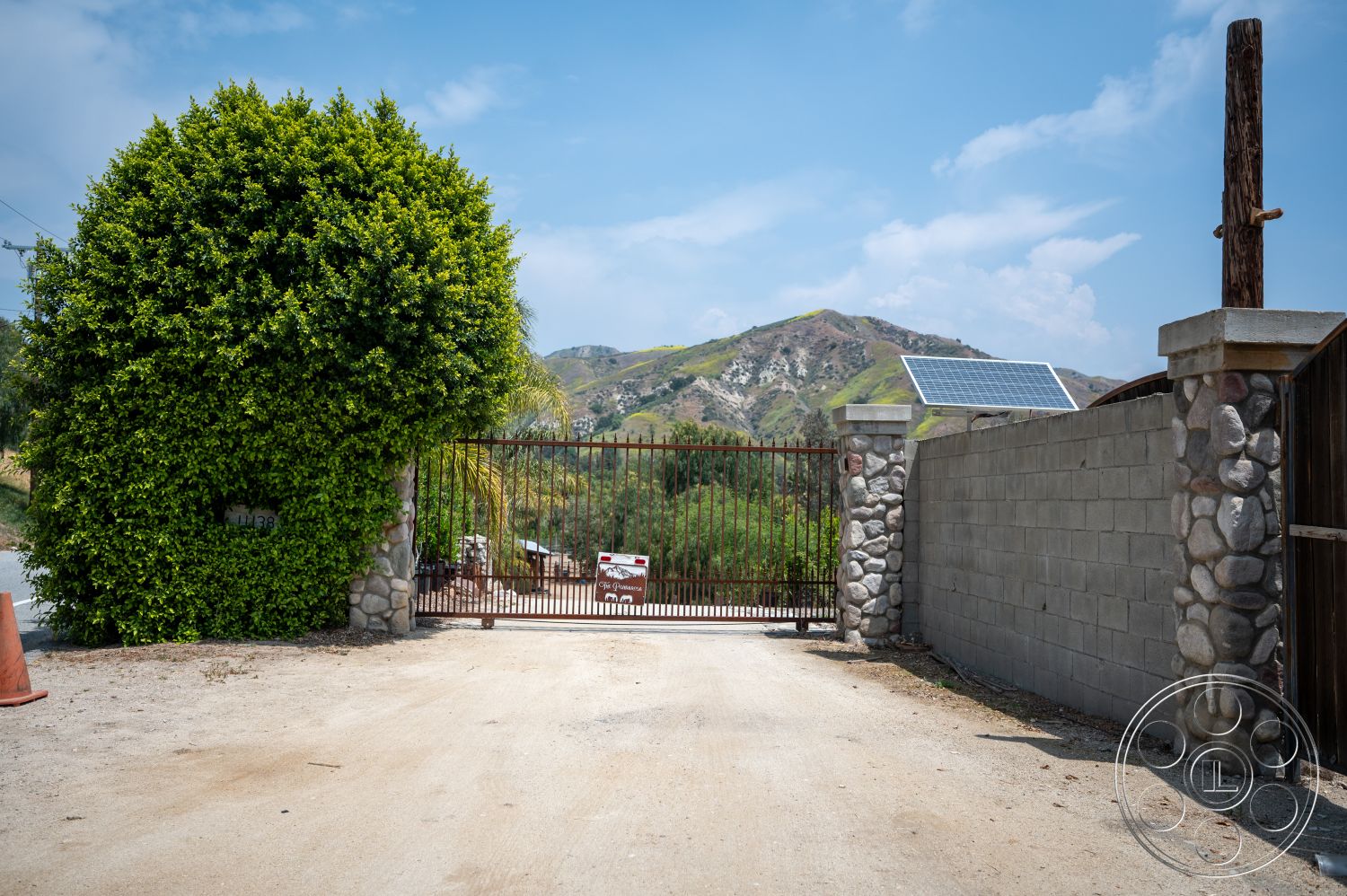 Ranch 6 - exterior, mountain view, green hedges, dirt driveway, concrete block wall, wrought iron gate, stone gate pillars, rustic wooden post, solar panel on gate