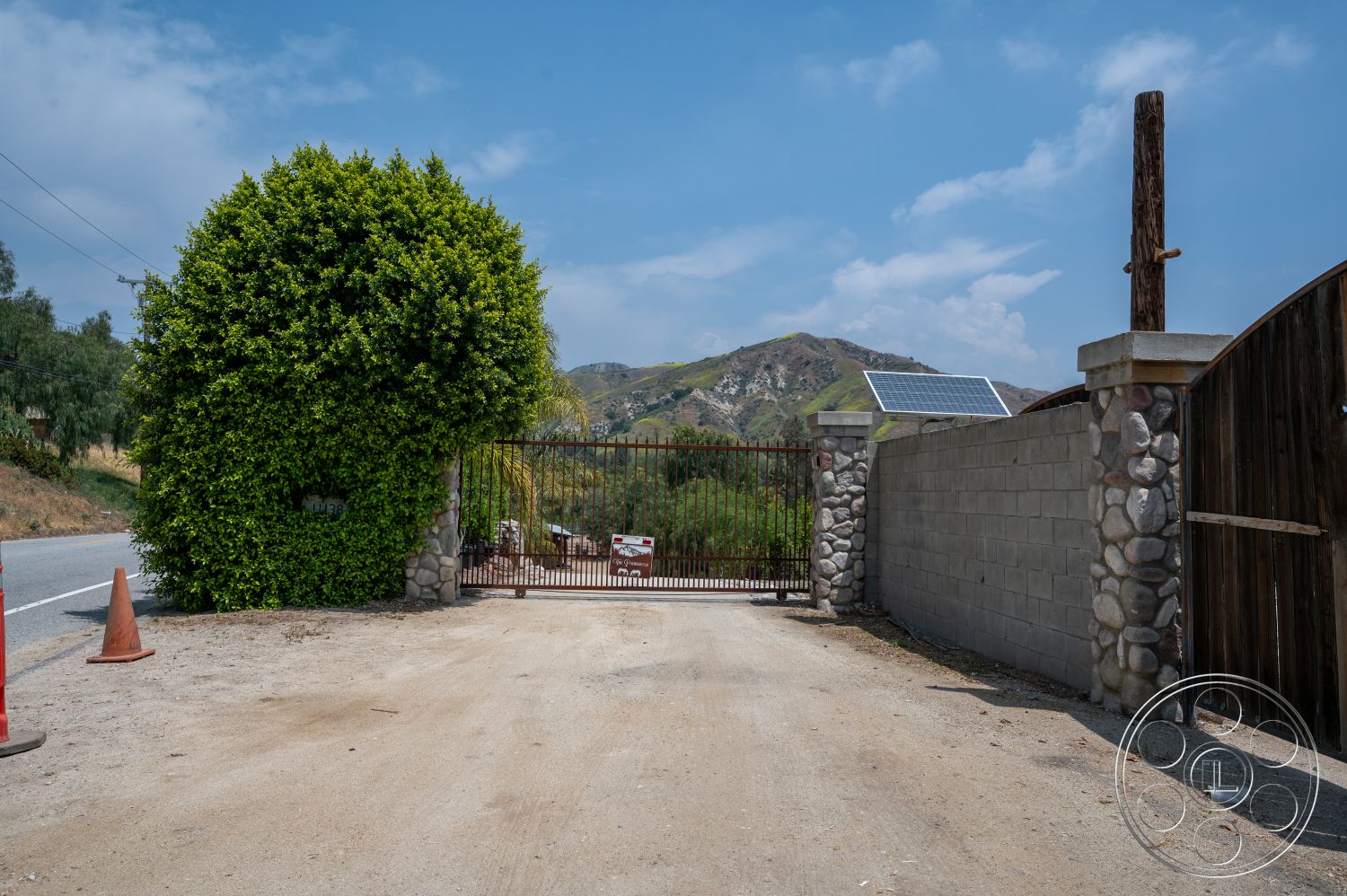 Ranch 6 - exterior, mountain view, rural setting, concrete block wall, solar panel installation, stone entrance gate, wooden gate structure, pebble stone wall, paved dirt driveway, large hedge