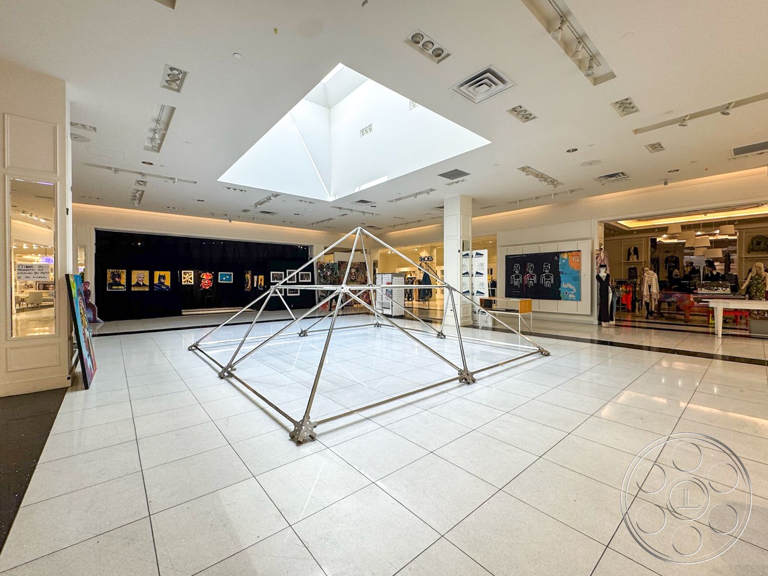 Beverly Gallery - interior space, polished stone floor, bright fluorescent lighting, open floor layout, white ceiling with recessed lighting, large glass skylight, metal geometric sculpture, black wall with framed artwork, shopping mall environment, display area