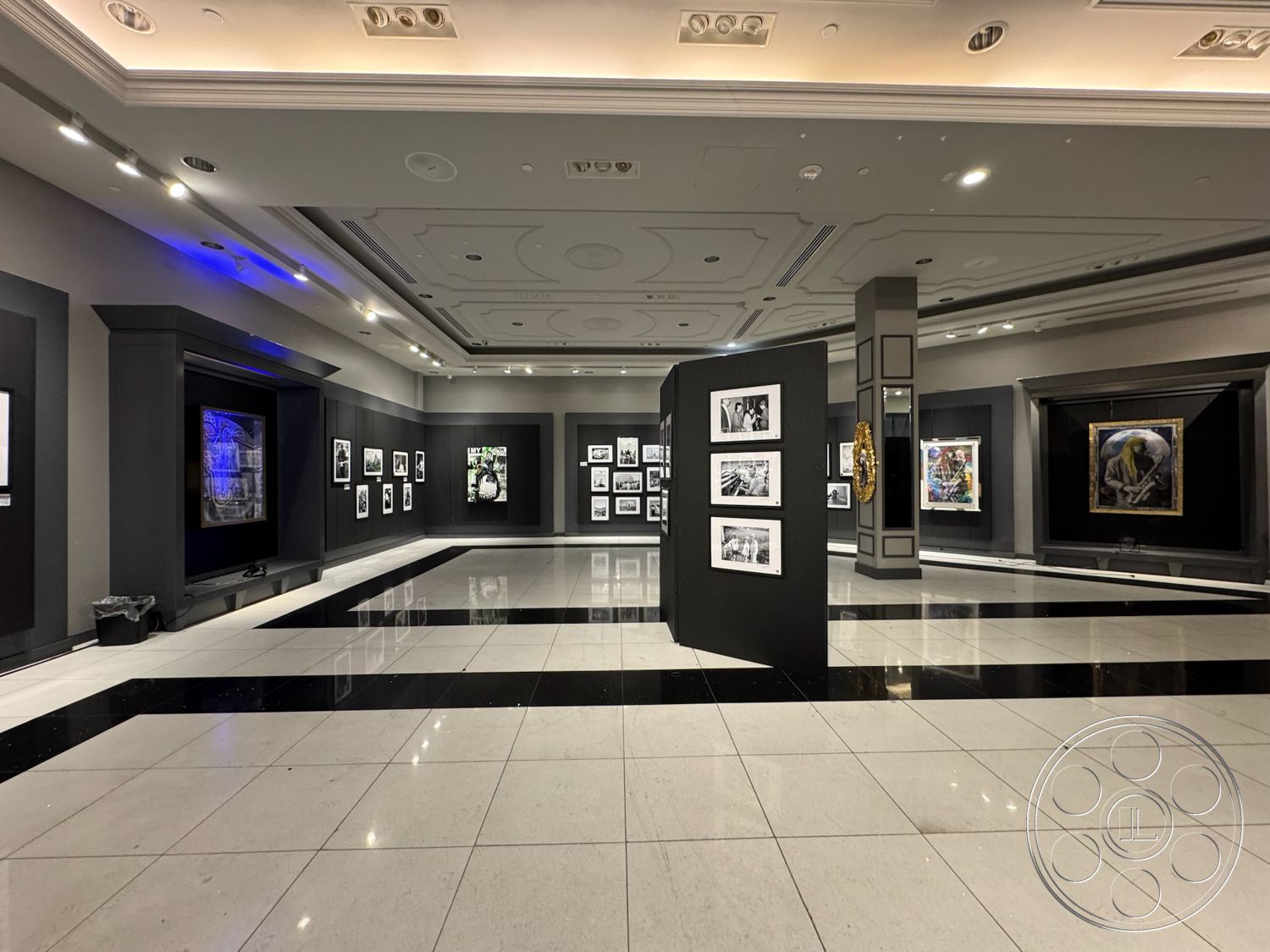 Beverly Gallery - polished marble flooring, black accent wall, exposed ceiling details, interior art gallery, black and white tile pattern flooring, shadowed wall with artwork displays, gallery lighting fixtures, modern exhibition display panels, framed photographs on walls, decorative gold wall art