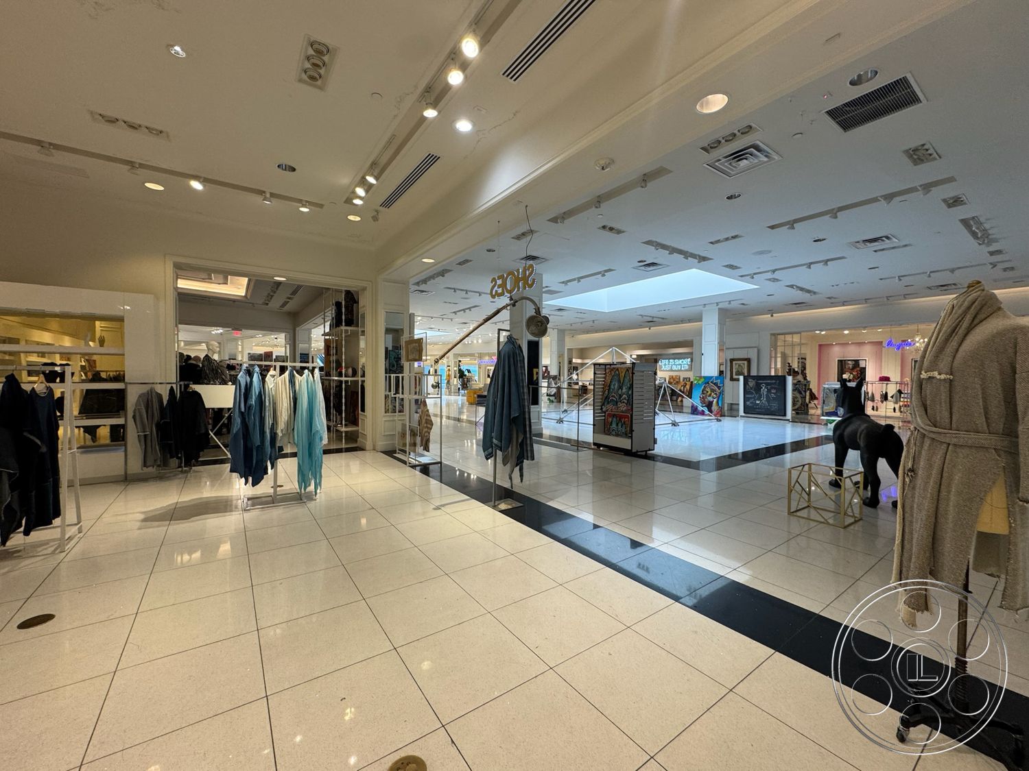 Beverly Gallery - open layout, glossy tile flooring, glass display cases, suspended ceiling with recessed lighting, mannequin display, brightly lit environment, art display walls, modern retail space, white ceiling tiles, clothing racks with hanging garments, prominent skylight, black and white tiled floor pattern