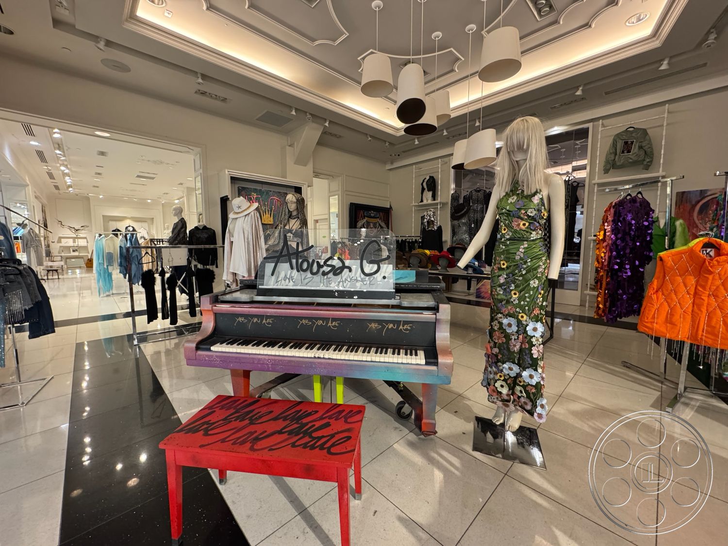 Beverly Gallery - polished marble flooring, glass partition walls, high ceiling with recessed lighting, pendant light fixtures, decorative ceiling molding, contemporary ceiling details, glossy black and white tiled floors, colorful painted upright piano, red painted piano bench with text, mannequin displaying floral dress, retail clothing display racks, modern clothing boutique design, brightly lit retail space, fashion merchandise display