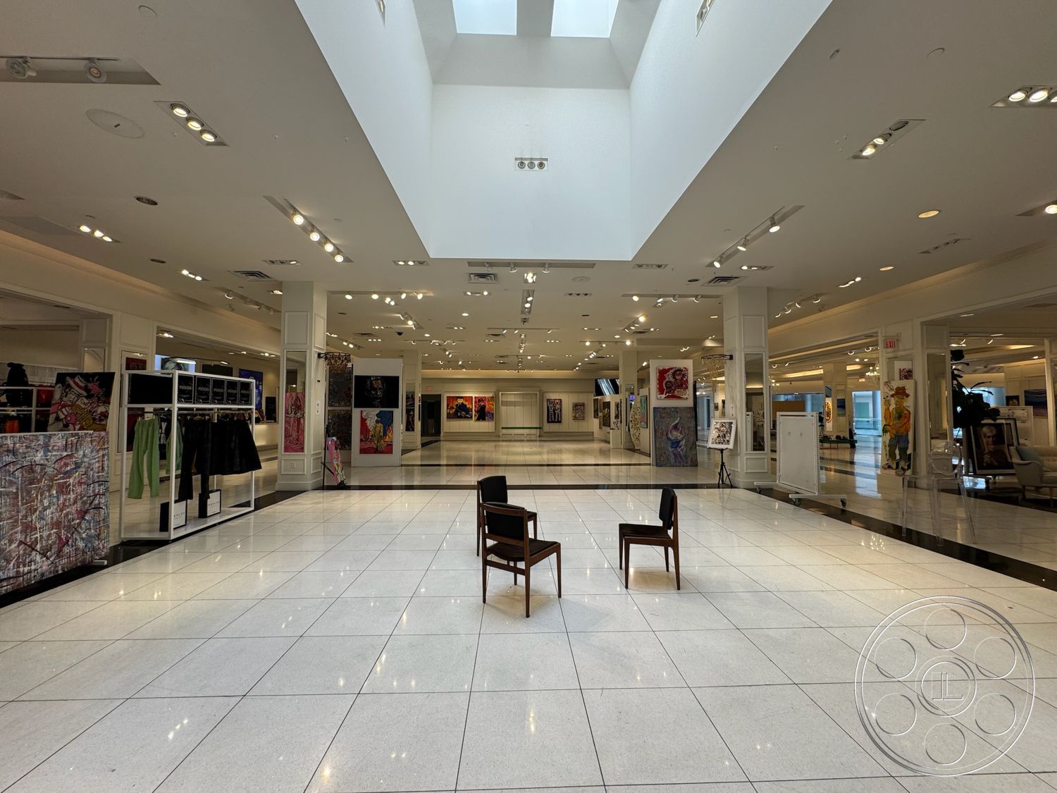 Beverly Gallery - neutral color palette, bright ambient lighting, two wooden chairs, open floor plan layout, white painted walls, minimalist furniture design, high ceiling with skylights, modern recessed lighting, glossy white tile flooring, displays for clothing and artwork, large wall art installations, broad hallway space