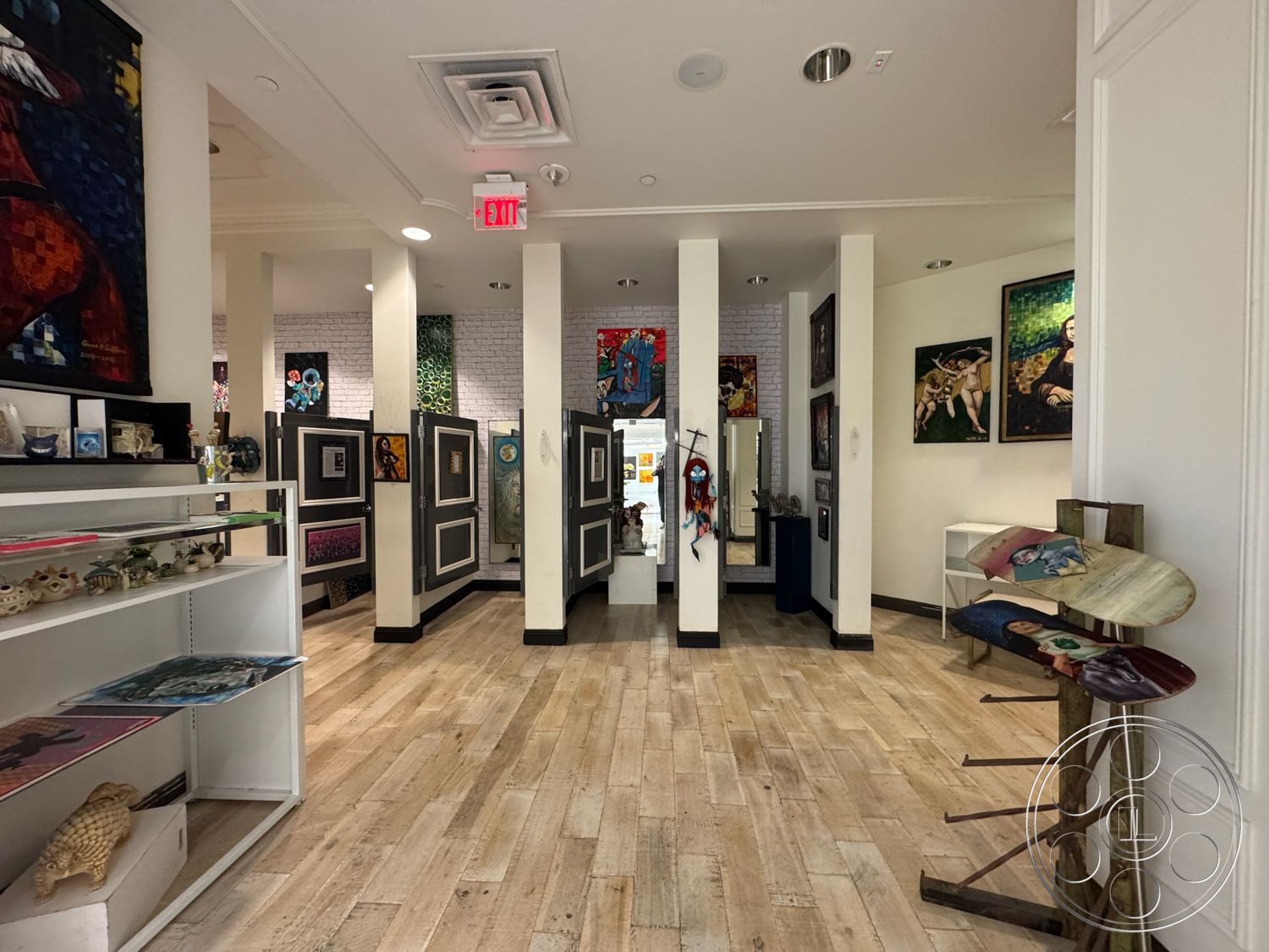 Beverly Gallery - open floor plan, track lighting, art gallery setting, framed art on walls, white painted walls, built-in shelving units, exposed brick accent wall, ceiling tiles, wood plank flooring, decorative artwork displays, black baseboards, colorful art installations