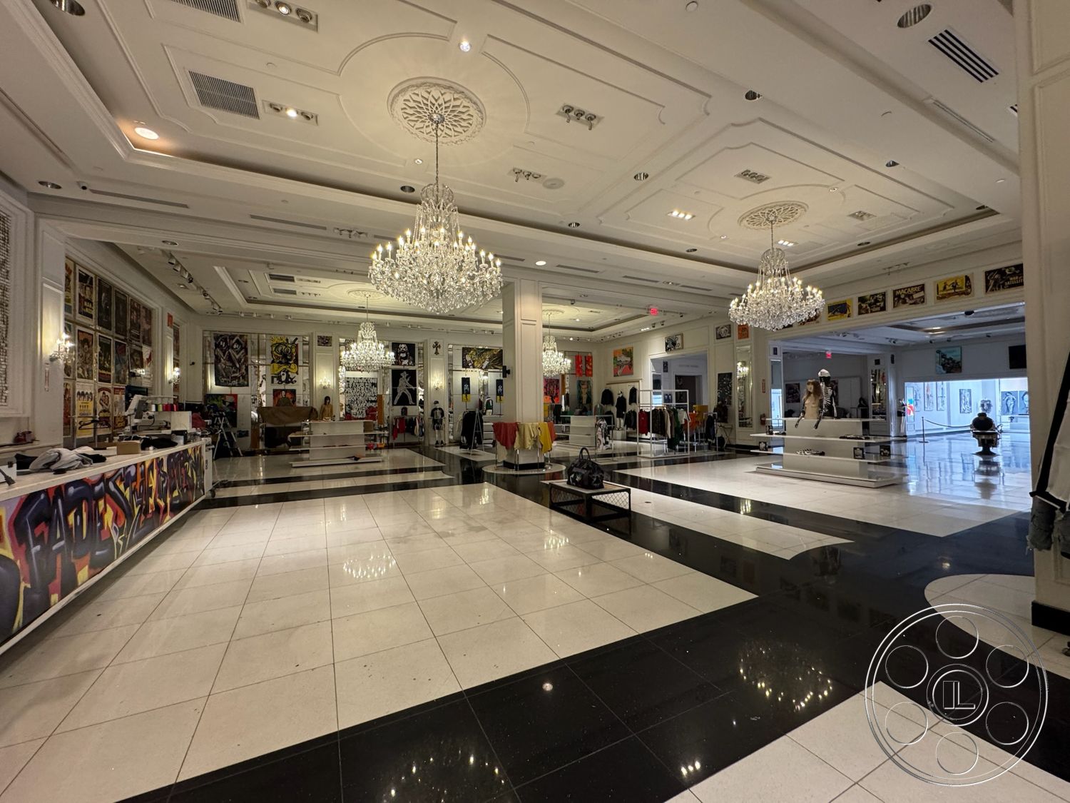 Beverly Gallery - chandelier lighting, bright natural light from large windows, black tile accents, white coffered ceiling, black and white polished tile flooring, gallery-style wall art displays, elegant retail space, modern clothing display stands, decorative wall molding details, stylized graffiti artwork, square-shaped floor plan