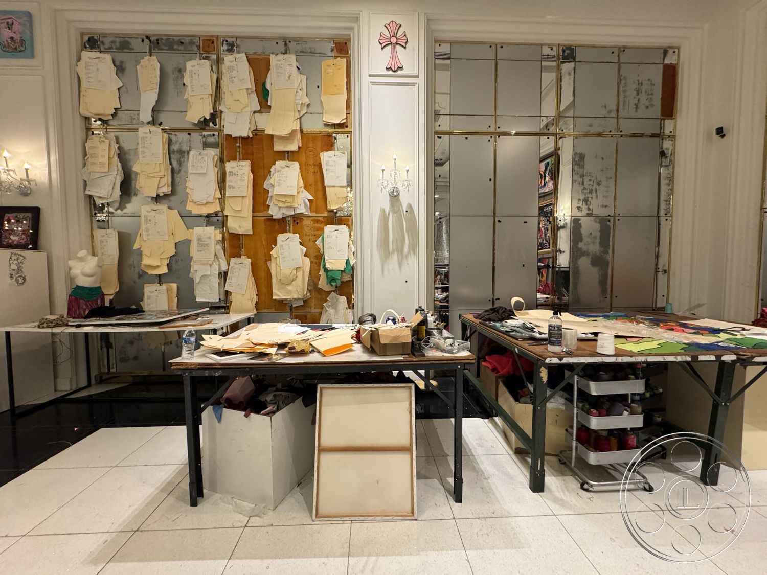 Beverly Gallery - lighting fixtures, decorative wall art, white plaster walls, black tile flooring, wooden table surface, creative studio environment, interior workspace, metal frame tables, organized cluttered workspace, mirror wall panels, design sketches on wall, hanging garment patterns, multi-color fabric swatches