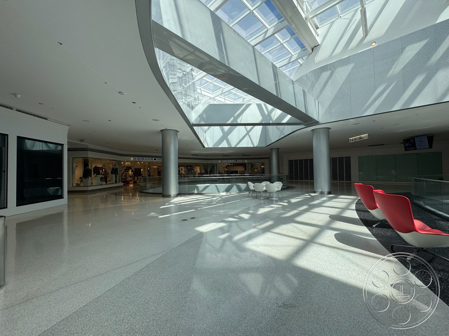 Beverly Gallery - neutral color palette, minimalist design elements, polished terrazzo flooring, floor-to-ceiling glass windows, curved glass skylight, open retail space, contemporary round seating area, stainless steel architectural columns, bright modern lighting