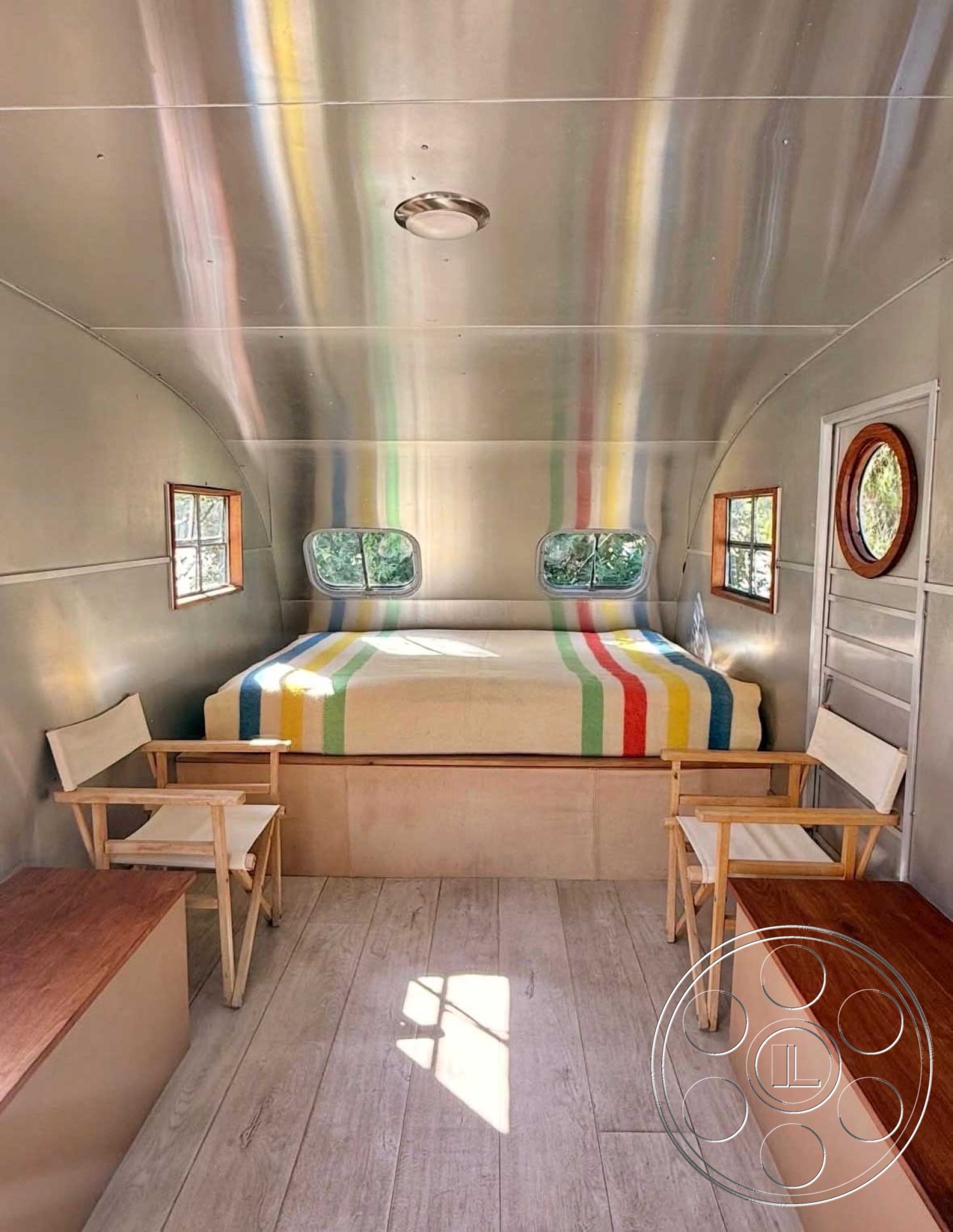 Zen Garden - minimalist design, natural wood accents, compact living space, wood plank flooring, aluminum interior walls, built-in bed platform, textured blanket with stripes, padded seating area, circular window details, square window frames, folding canvas chairs