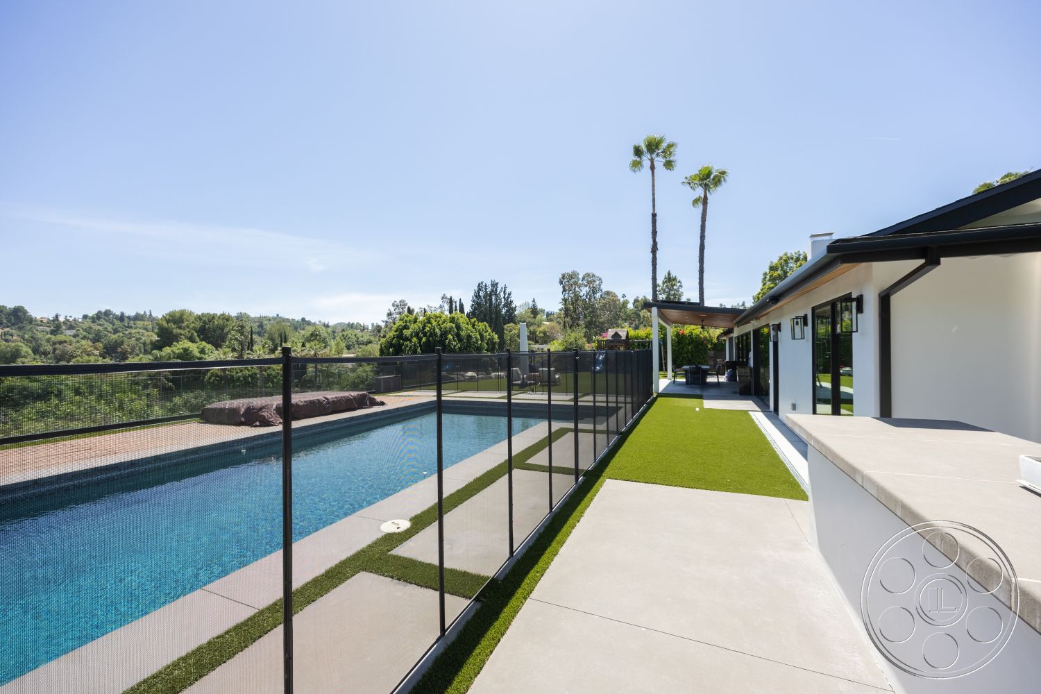 Modern 390 - exterior, large glass sliding doors, rectangular swimming pool, artificial grass landscaping, flat roof design, modern single-story home, concrete pool border, concrete patio floor, clear blue pool water, black mesh pool safety fence, palm trees surrounding property, lush green hillside view
