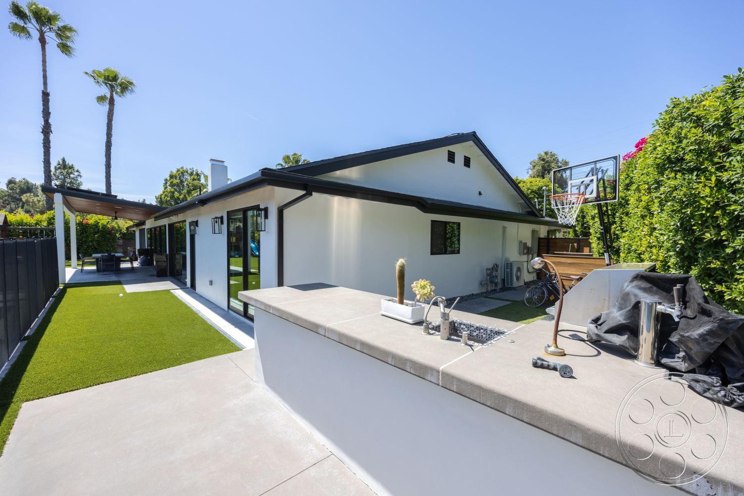 Modern 390 - exterior, white stucco walls, large glass sliding doors, gabled roof, black metal fence, palm trees in background, flat roof design, modern outdoor seating area, concrete patio floor, landscaped garden borders, synthetic grass lawn, outdoor basketball hoop, concrete counter with built-in sink