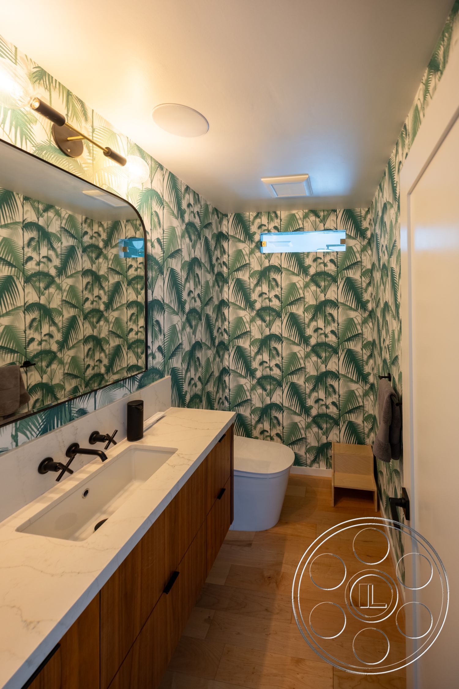 Modern 390 - round mirror, green floral wallpaper, neutral wall color, textured ceiling, white marble countertop, white toilet, small wooden stool, glass block window, wooden bathroom vanity, modern wall-mounted lighting, black faucet fixtures, pine hardwood flooring
