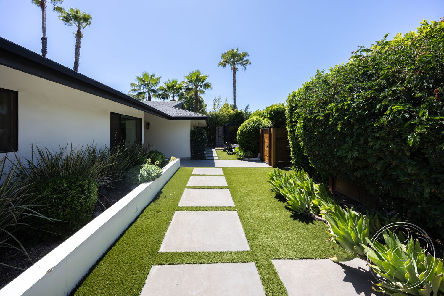 Modern 390 - exterior, outdoor living space, modern architectural style, tropical palm trees, flat roof design, white painted walls, landscaped garden beds, artificial grass lawn, surrounding hedge landscaping, square concrete stepping stones, wood privacy fence