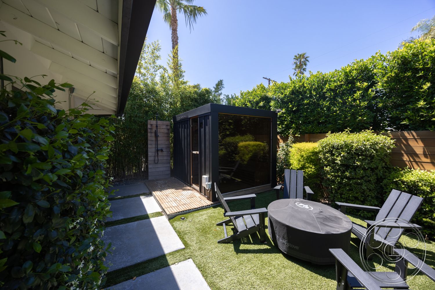 Modern 390 - exterior, outdoor seating area, adirondack chairs, lush green hedges, wooden privacy fence, wooden deck flooring, modern minimalist design, palm tree landscaping, open sky view, artificial grass lawn, concrete patio tiles, round fire pit table, black modular garden shed, paved walkway stones