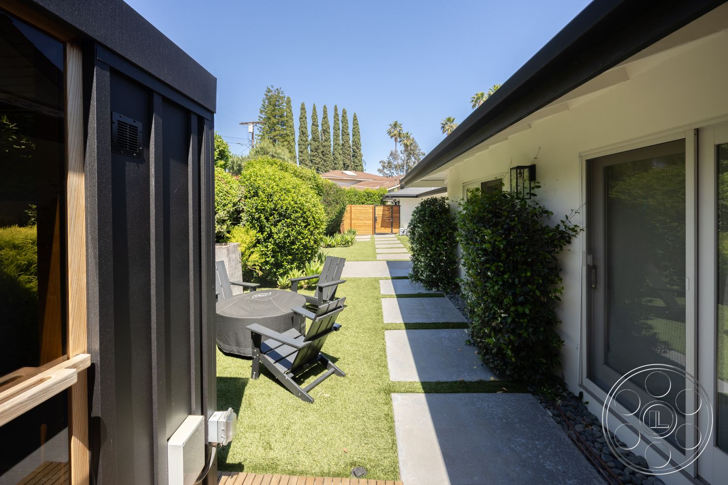 Modern 390 - exterior, sliding glass doors, concrete pathway, outdoor dining area, grass lawn, clear blue sky, tall cypress trees, wooden outdoor seating, modern house facade, paved stepping stones, black metal shed, landscape planting, green privacy hedge