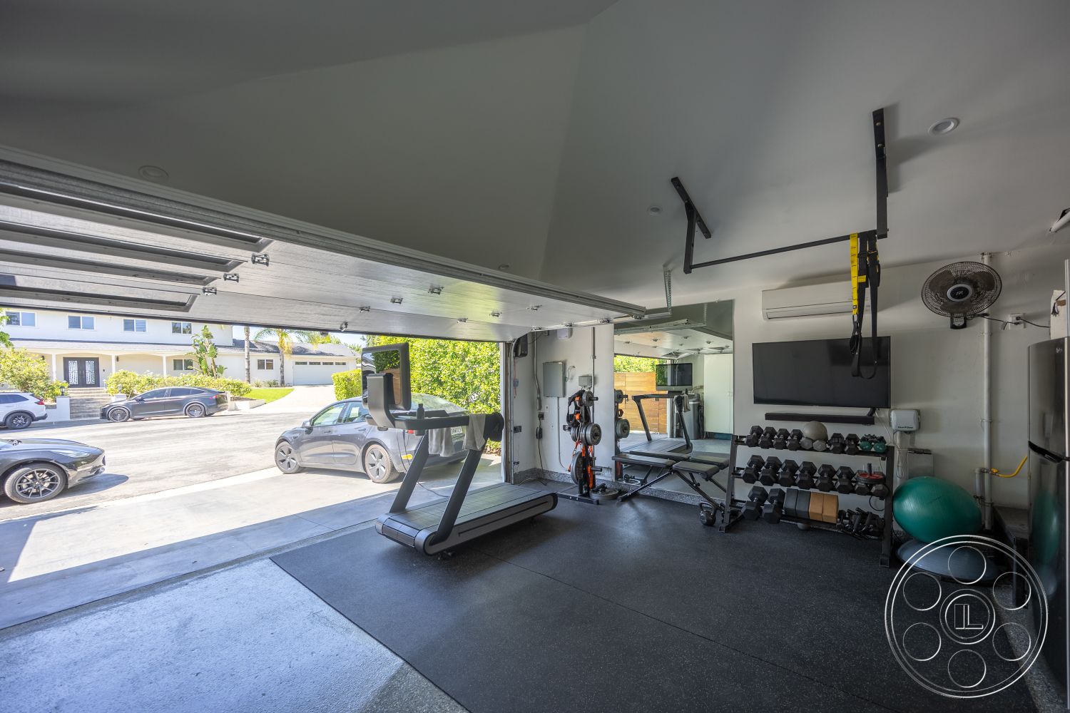 Modern 390 - wall-mounted television, rubber flooring, open space layout, mirror wall treatment, interior gym space, lifting weights area, treadmill apparatus, ceiling with recessed lighting, hanging suspension straps, exposed ventilation ducts, garage door opening, fitness equipment storage, natural light from opening
