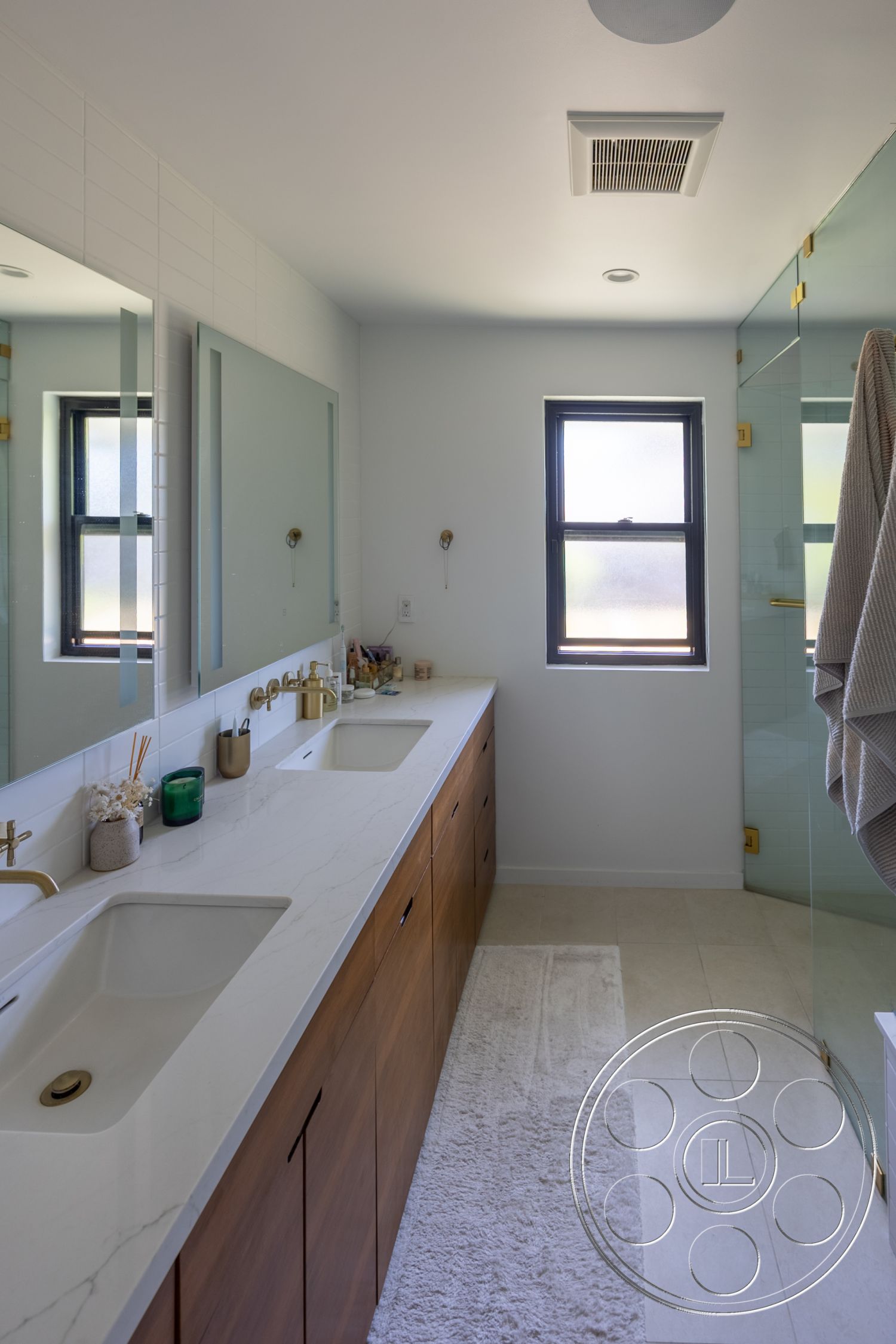 Modern 390 - glass shower enclosure, gold fixtures, rectangular mirrors, natural light from window, minimalist bathroom design, light gray tile flooring, white tiled backsplash, white marble bathroom countertops, dark wood vanity cabinets, white fluffy bath mat, modern sink design, brushed gold hardware