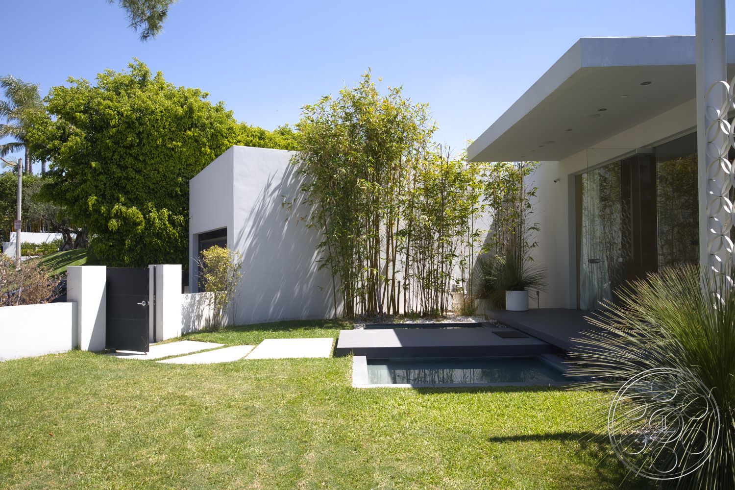 Modern 525 - exterior, white stucco walls, glass sliding doors, black metal gate, rectangular pool, concrete stepping stones, stone pool deck, landscaped garden area, minimalist architectural style, concrete patio area, flat-roofed modern home, lush green bamboo privacy screen, curved landscaping bed