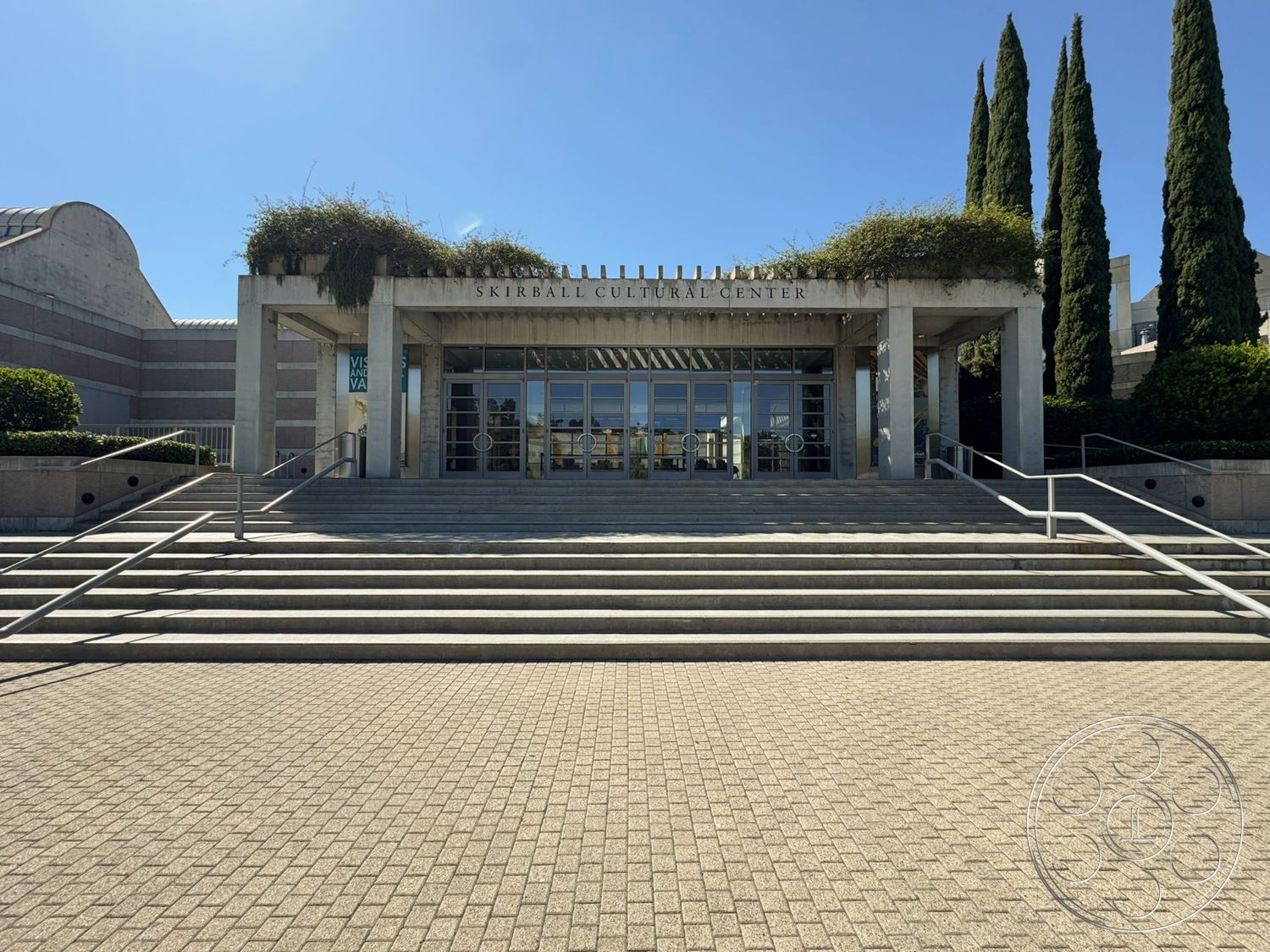 Museum 2 - exterior, concrete steps, cypress trees, landscaped garden beds, flat roof structure, metal handrails, glass entrance doors, stucco wall finishes, brick paver walkway, horizontal banded facade, open plaza space